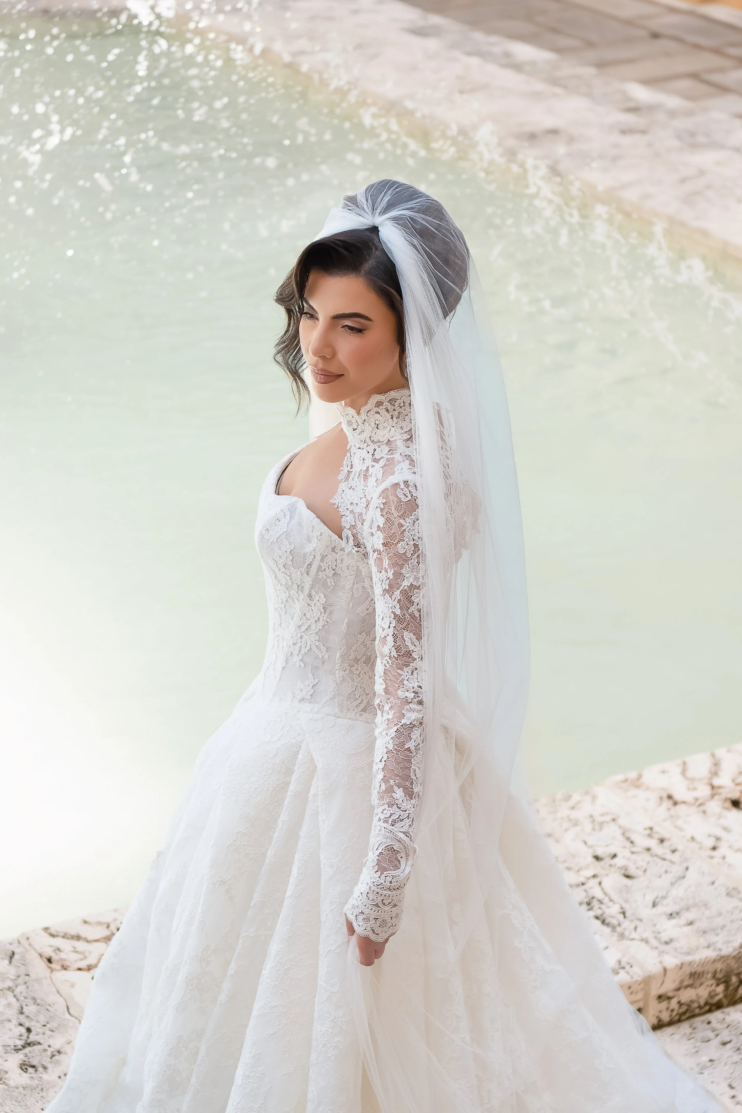 A woman in a white wedding dress with lace detail and a sheer veil, standing by a body of water with sandy shoreline.