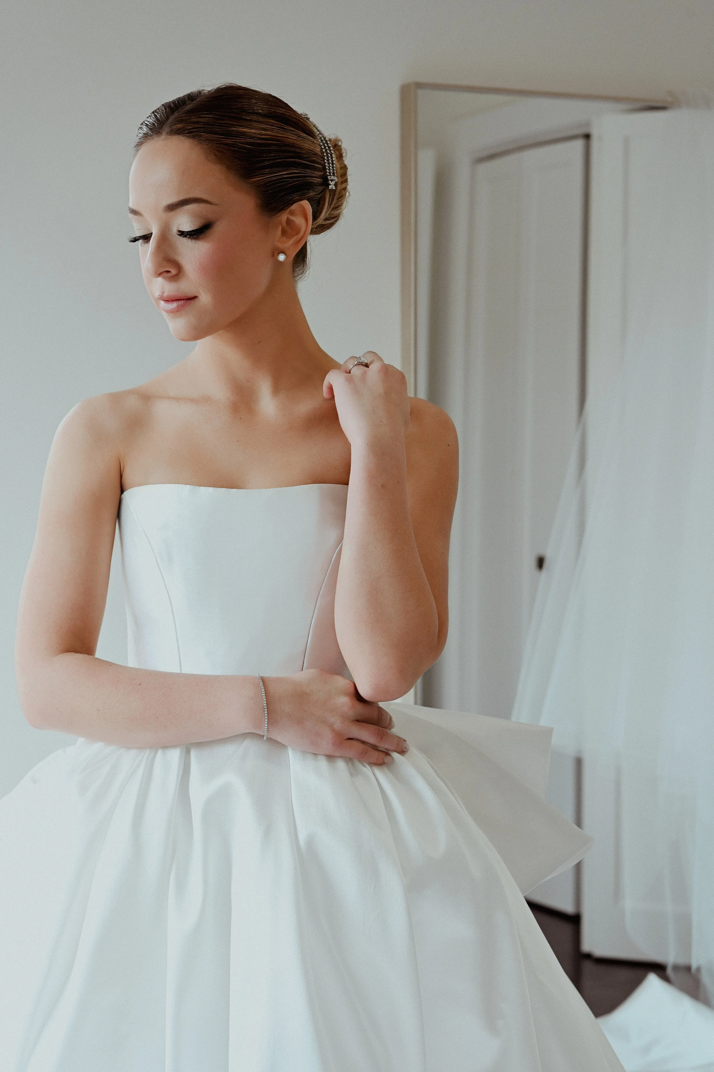 A woman in a white strapless dress, possibly a bride, is standing near a mirror with her eyes closed, looking downward, with her right hand touching her left shoulder. She has styled hair with a hair accessory and is wearing earrings, a ring, and a b