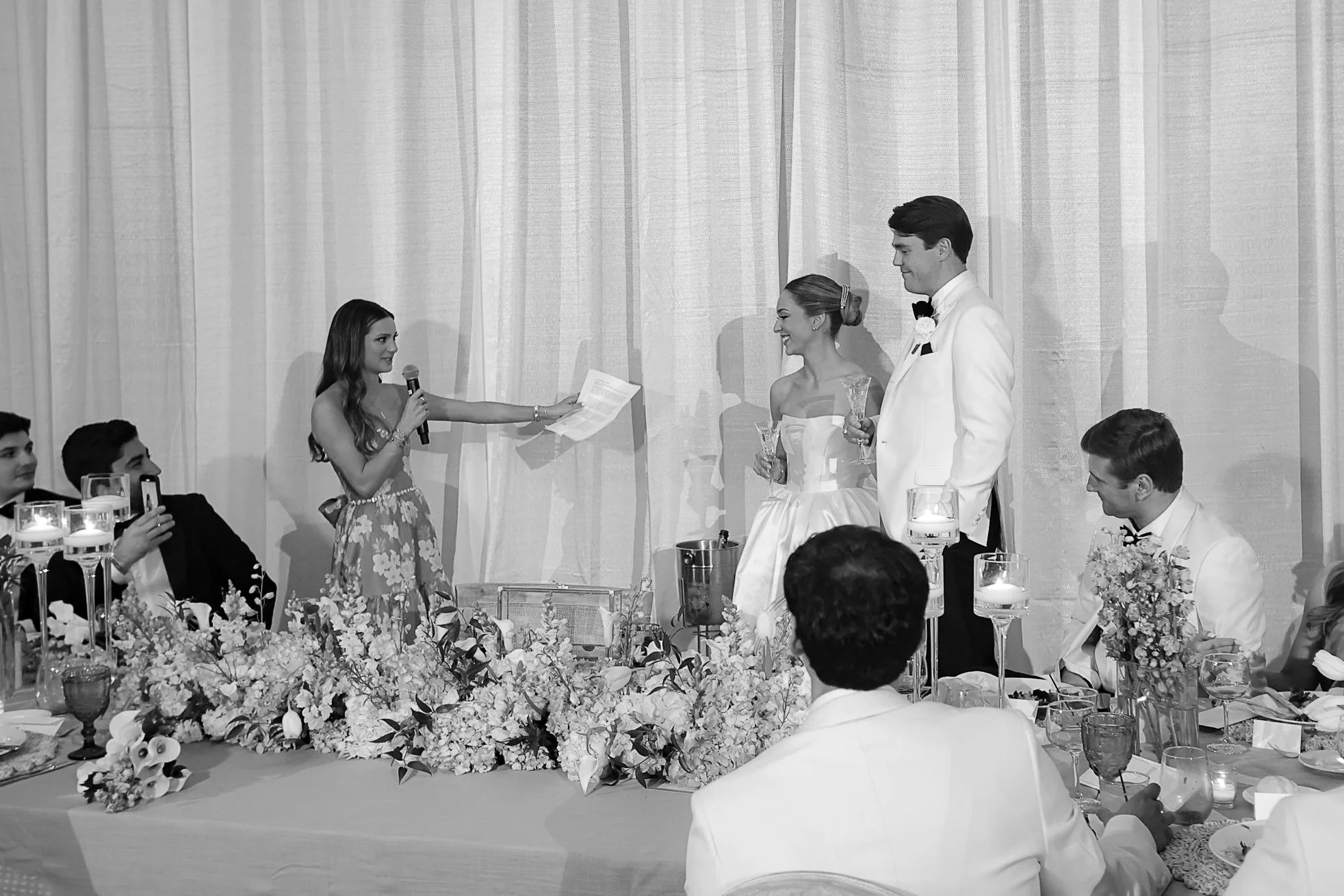 A black-and-white photo of a wedding reception with a bride and groom standing together, the bride holding a glass and the groom dressed in a tuxedo. A woman is speaking into a microphone, possibly giving a toast. Guests are seated at tables decorate