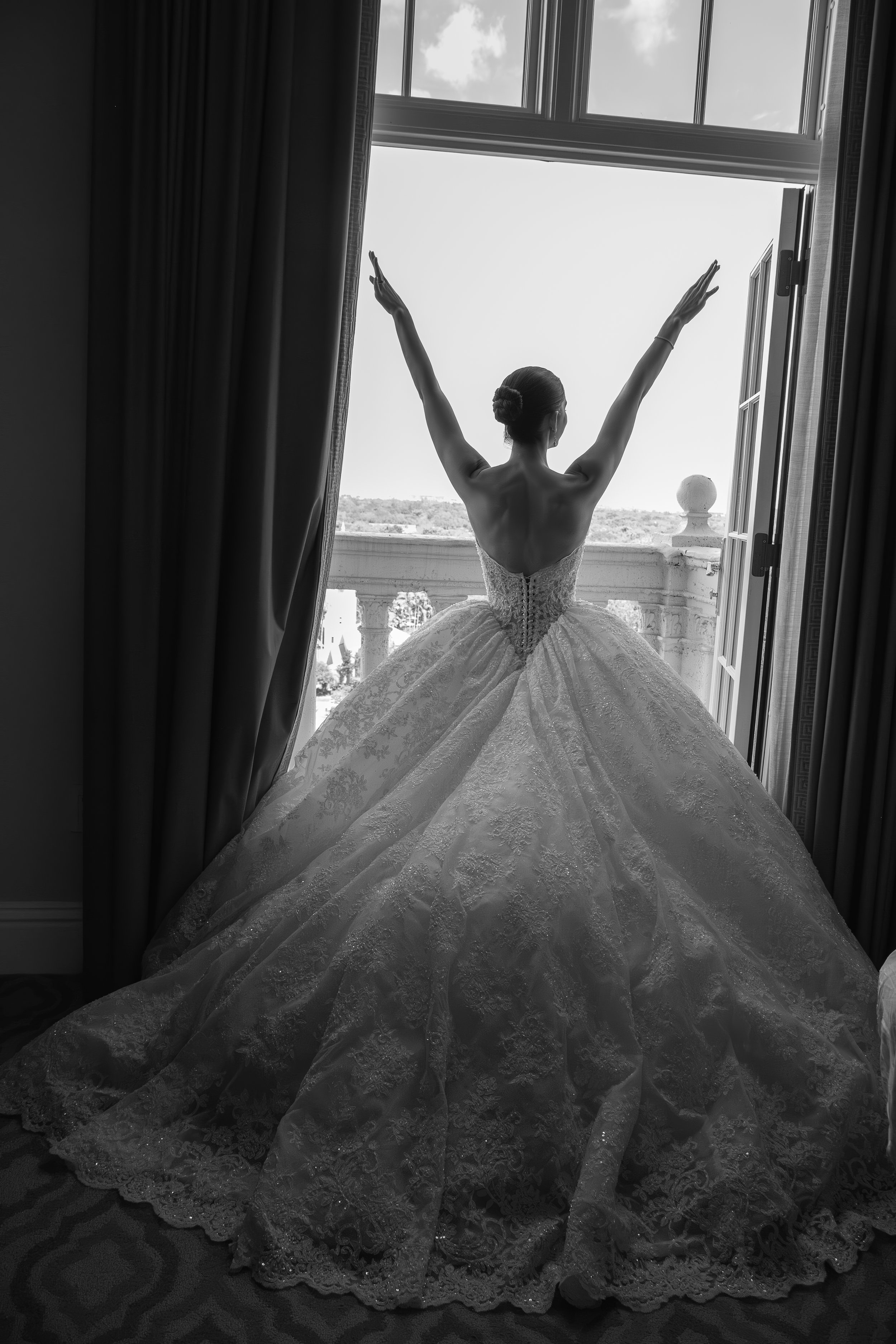 Black and white photo of a woman in a wedding dress standing on a balcony, with her arms raised, looking out the open window.