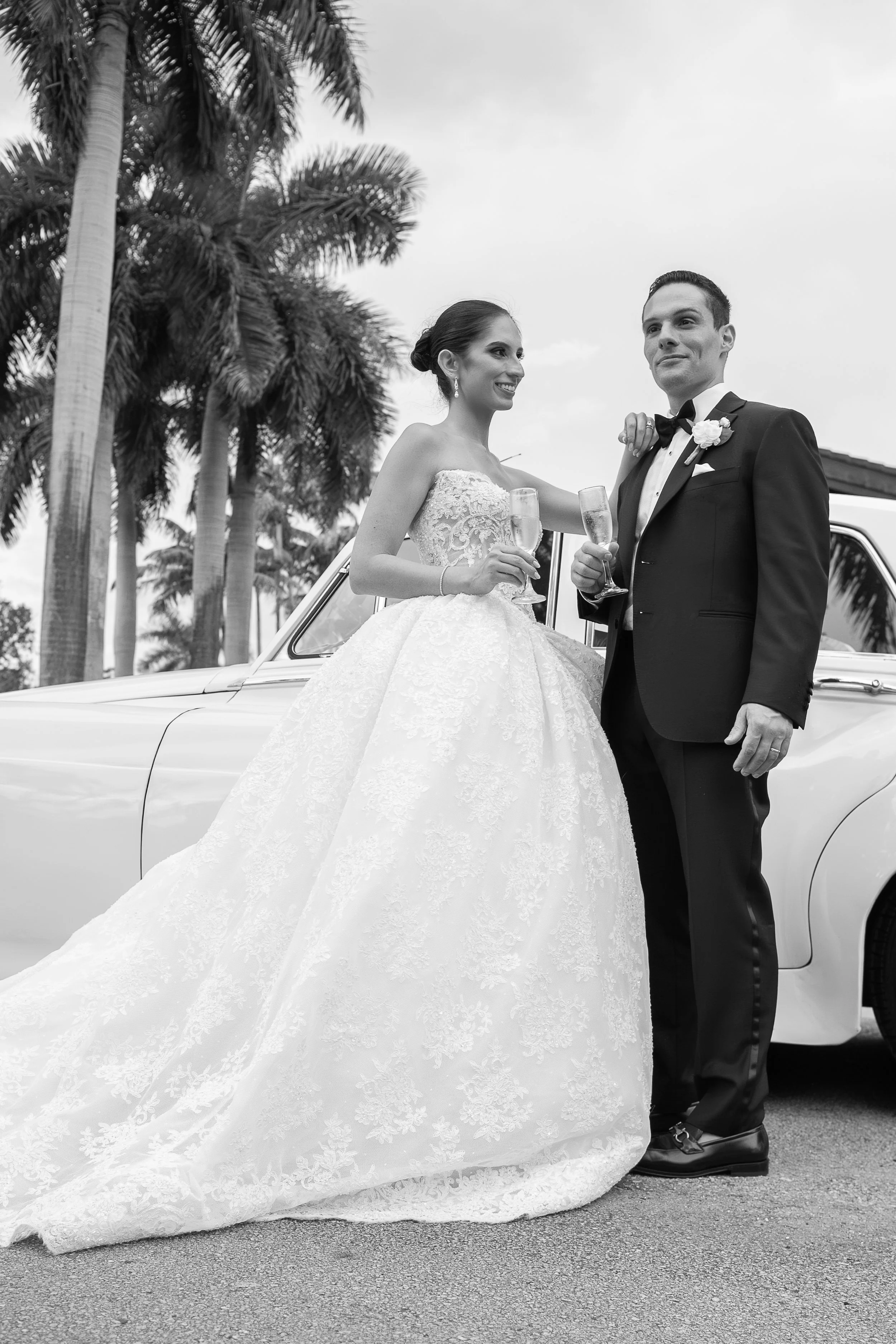 A black and white photo of a bride and groom standing next to a vintage car outdoors. The bride is in a strapless lace wedding gown holding a glass of champagne, and the groom is in a tuxedo with a bow tie also holding a champagne glass. There are pa