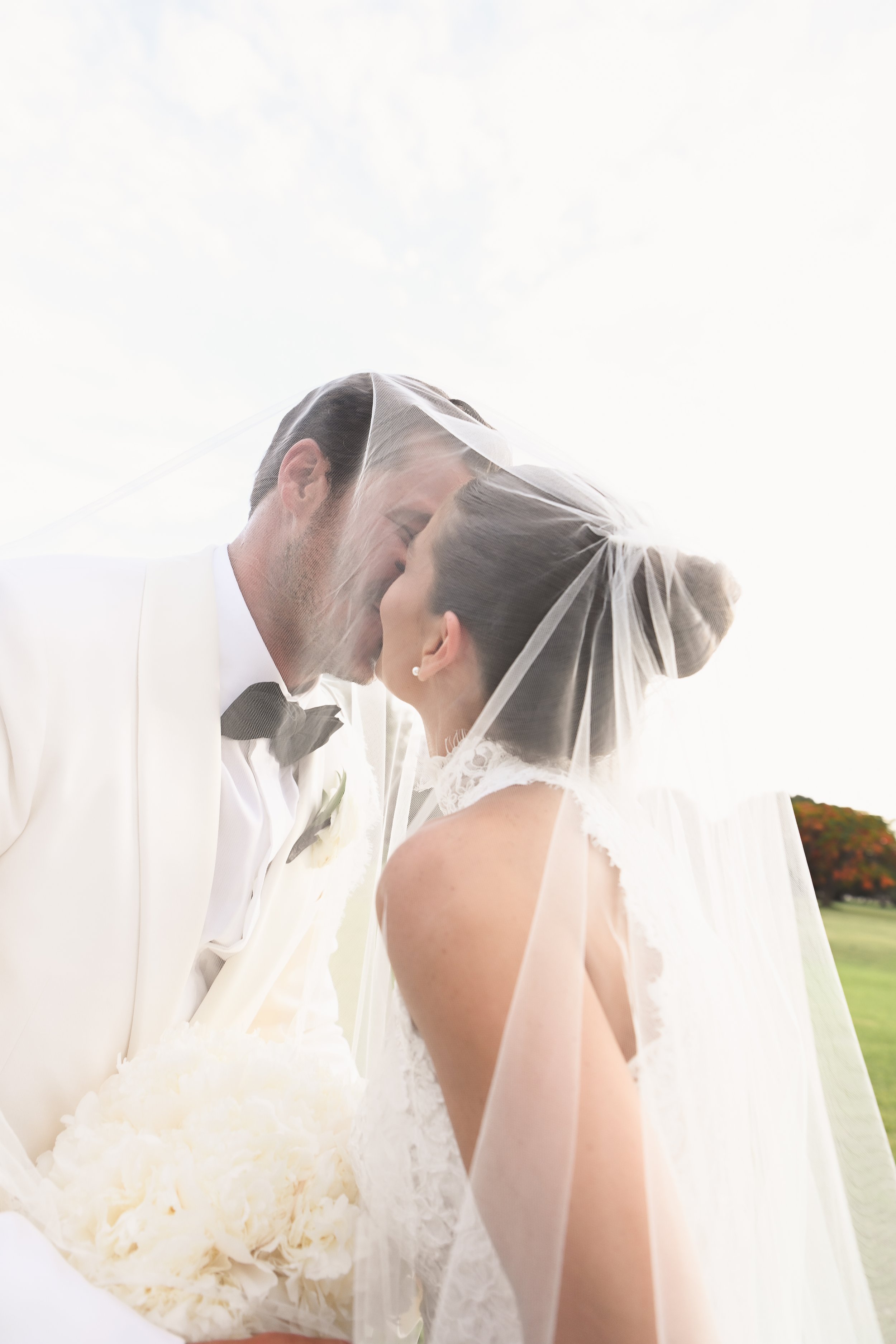 A bride and groom are sharing a kiss outdoors on a bright, overcast day. The groom is wearing a white tuxedo with a black bow tie, and the bride is in a white lace wedding dress with a veil. The groom holds a bouquet of white flowers.