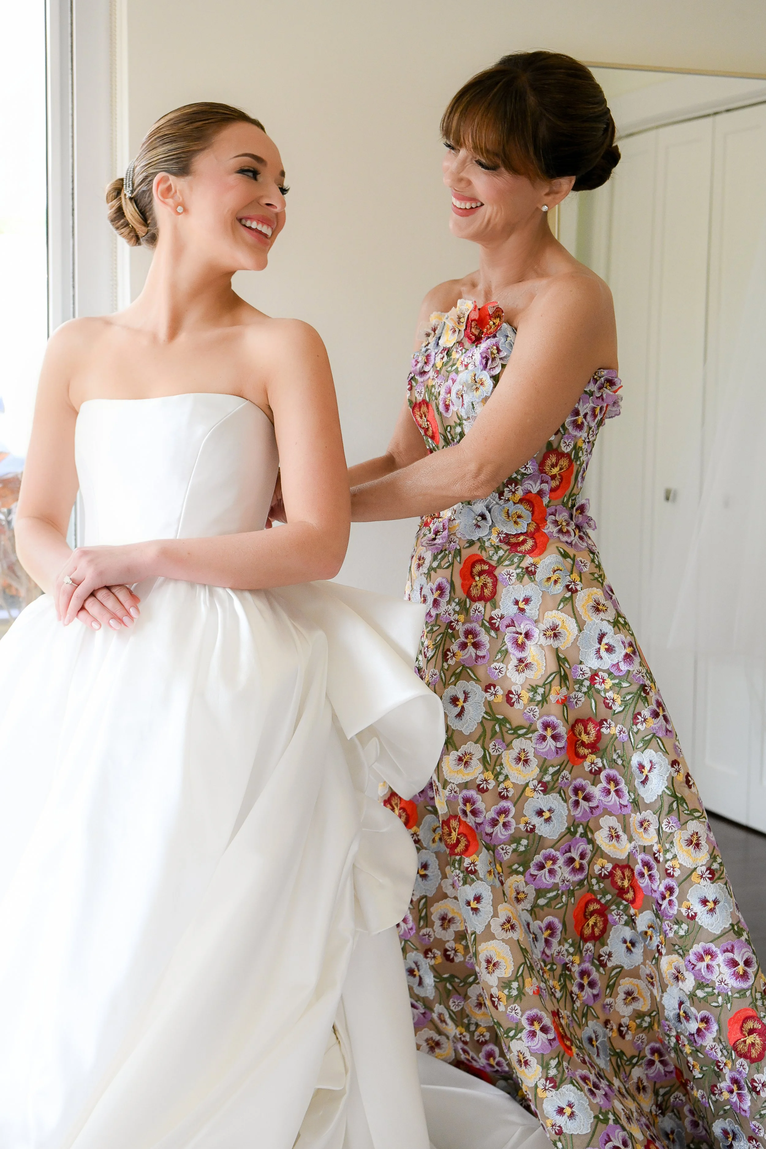A bride in a strapless white wedding gown smiling and looking at a woman in a floral dress, standing by a window in a bright room.