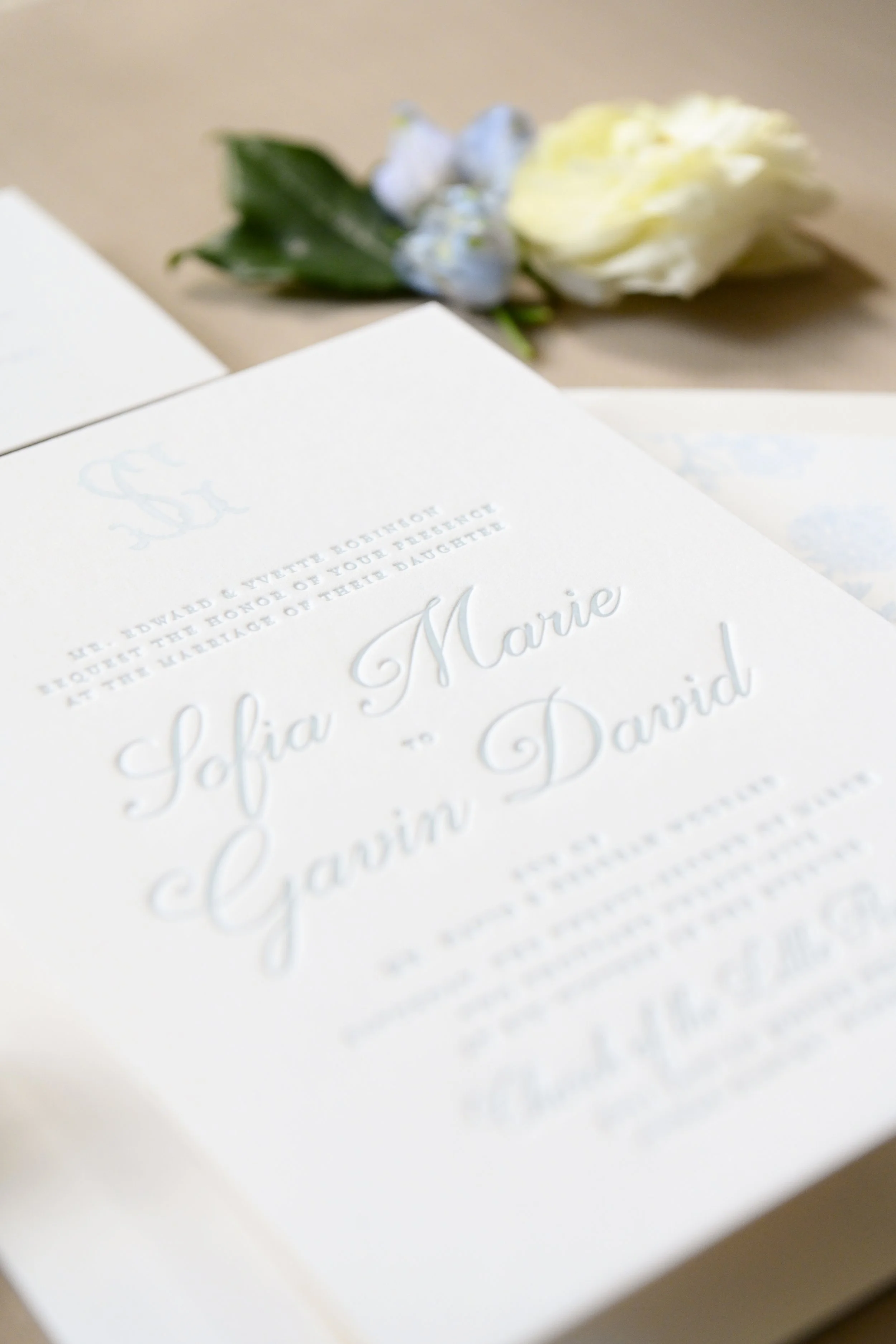 Close-up photo of a wedding invitation card with white embossed text and a small bouquet of white flowers in the background.