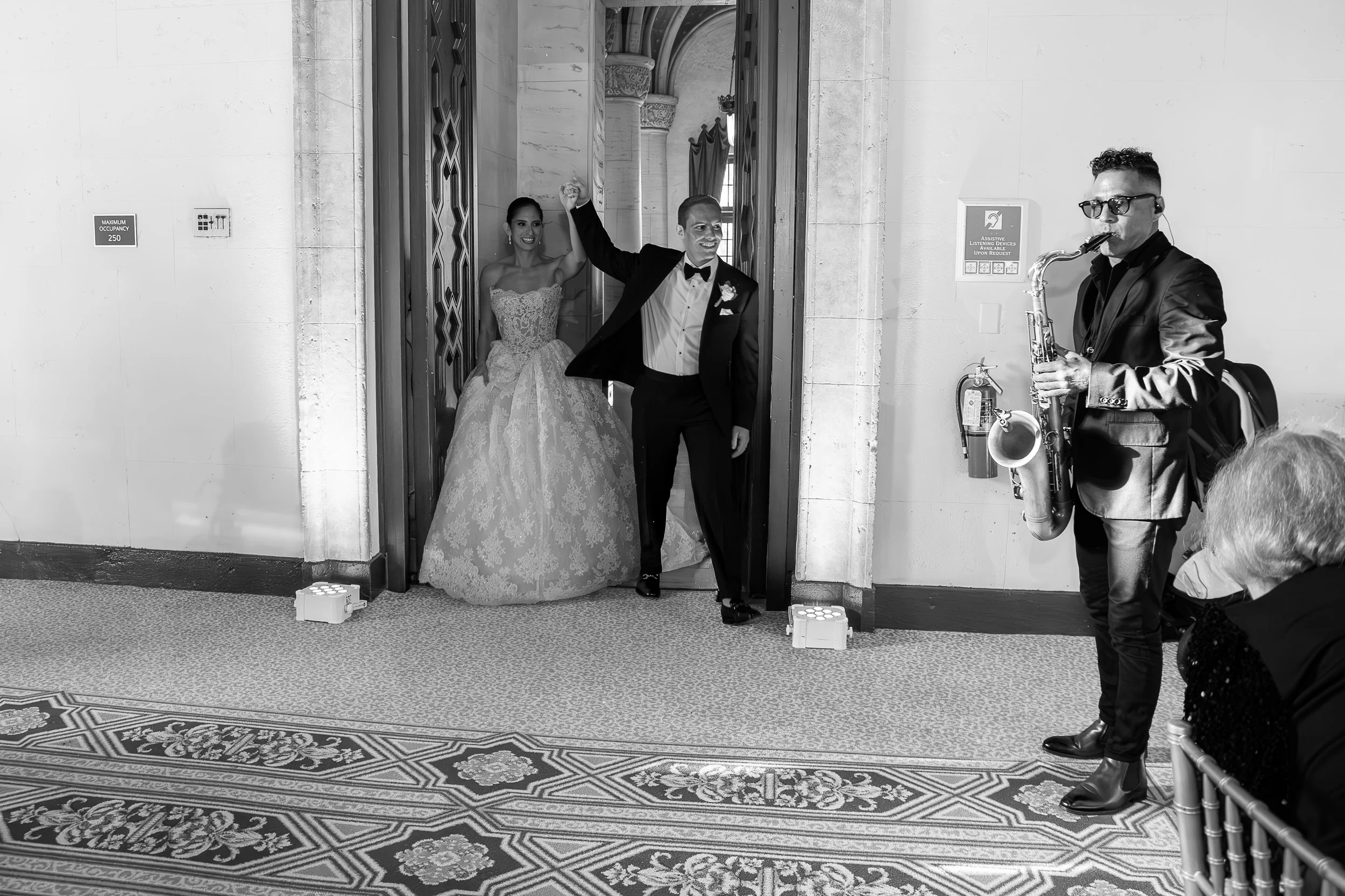 A bride and groom celebrating their wedding at an entrance with a musician playing saxophone to the right. The bride is in a wedding dress, and the groom is in a tuxedo, holding her hand as they walk out.