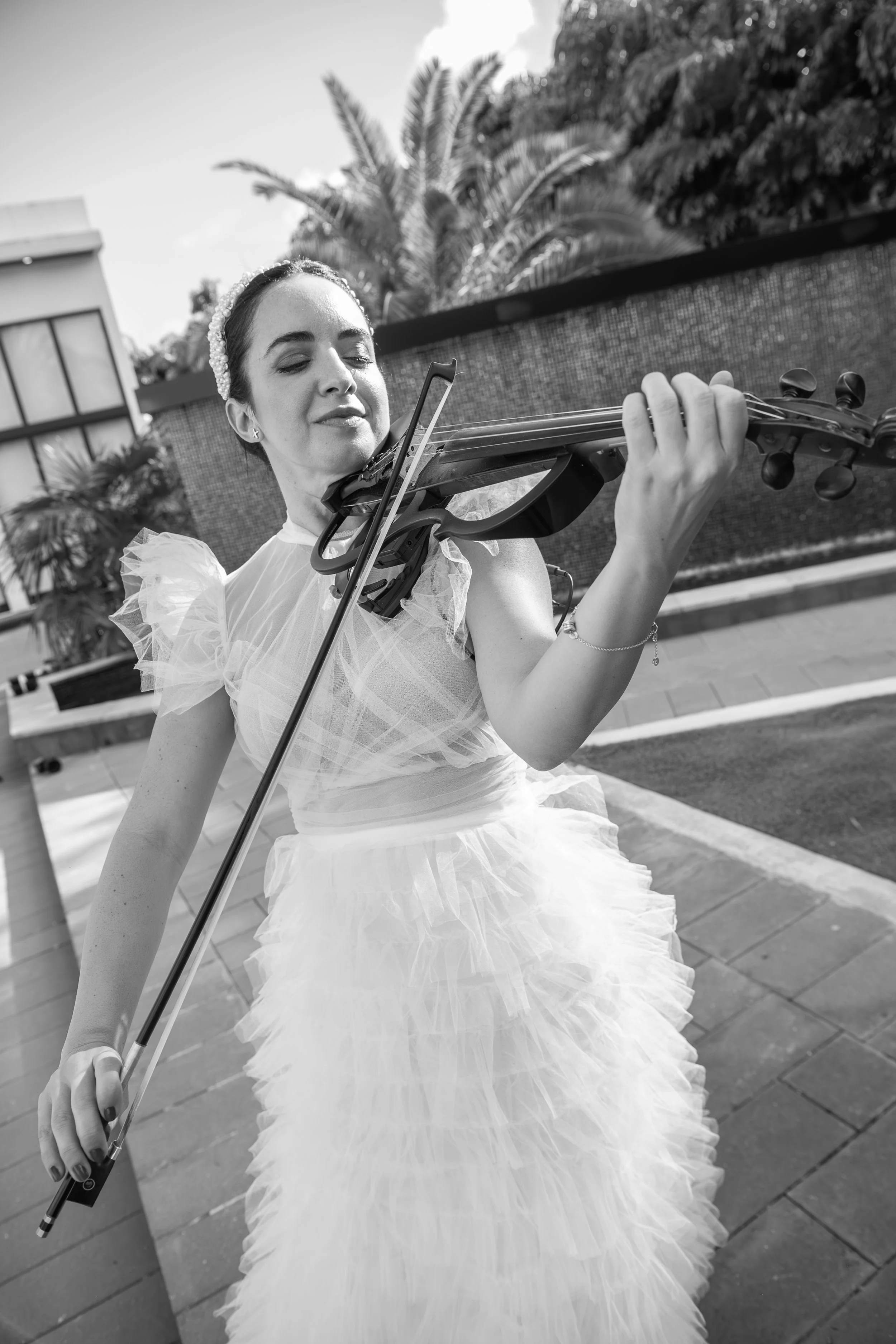 Woman in a wedding dress playing the violin outdoors, with trees and buildings in the background in black and white.