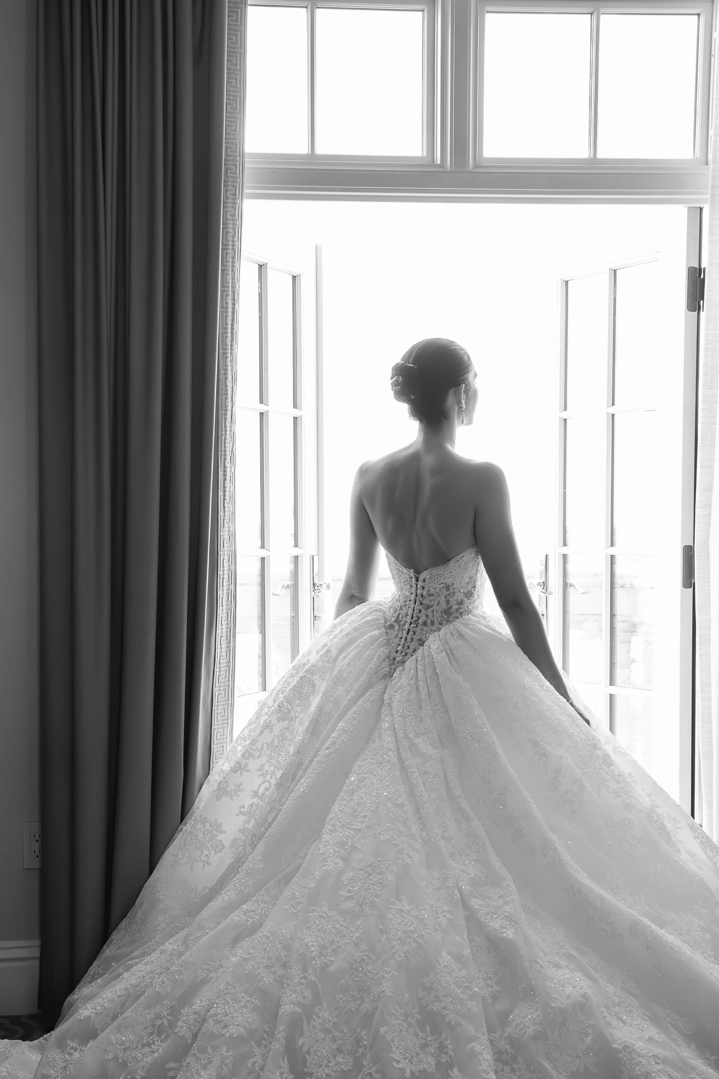 A woman in a wedding dress sitting by open windows, looking outside. The black and white photo captures her elegant hairstyle and detailed lace gown.