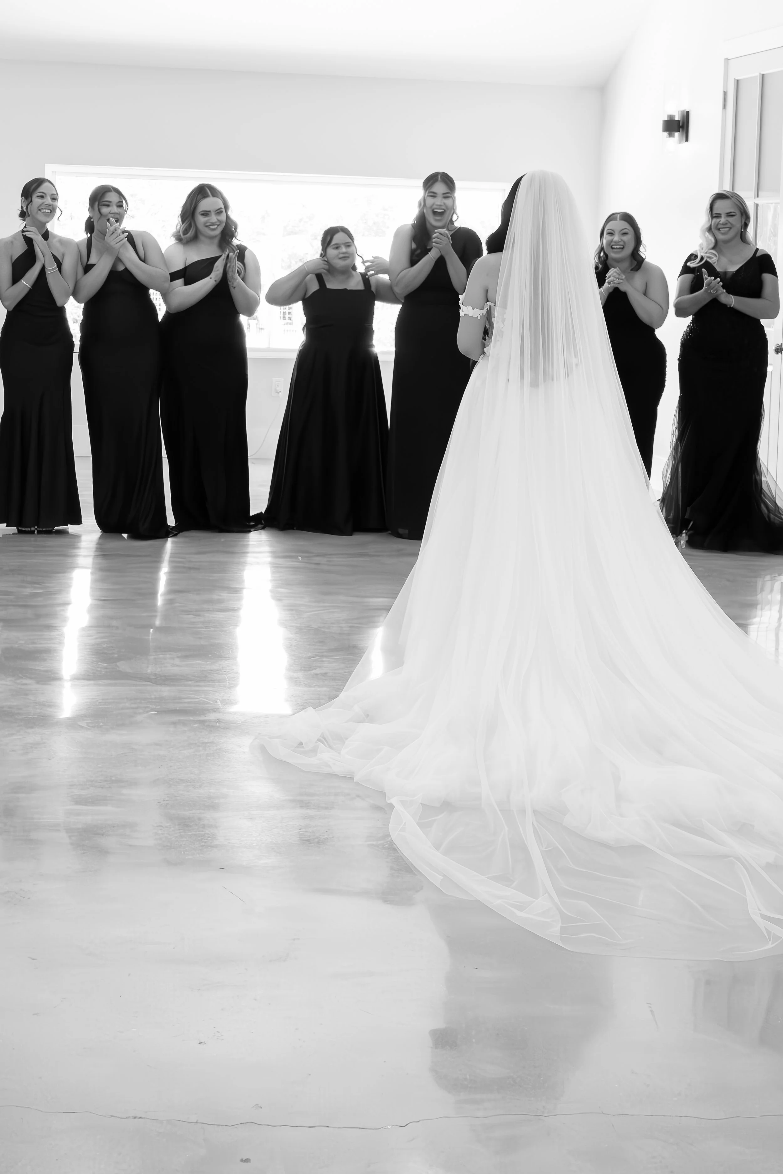 A bride in a wedding gown with a long train, facing a group of six women in matching black dresses, inside a well-lit room with wooden flooring and large windows.