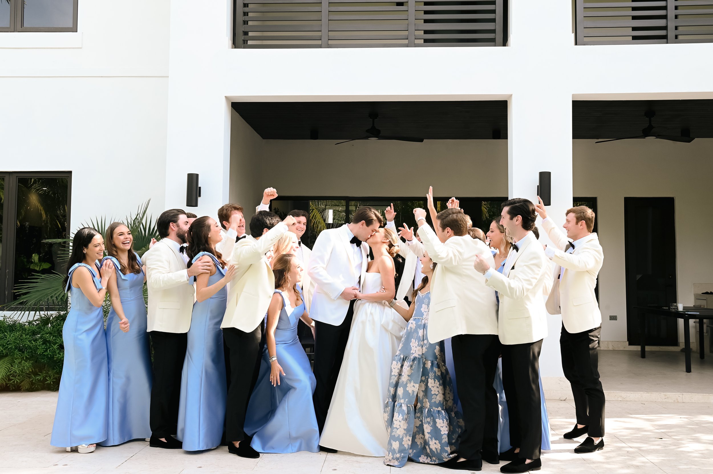 A group of people dressed in formal attire, including light-colored tuxedos and blue dresses, celebrating and kissing each other outside a modern building with white walls and a balcony.