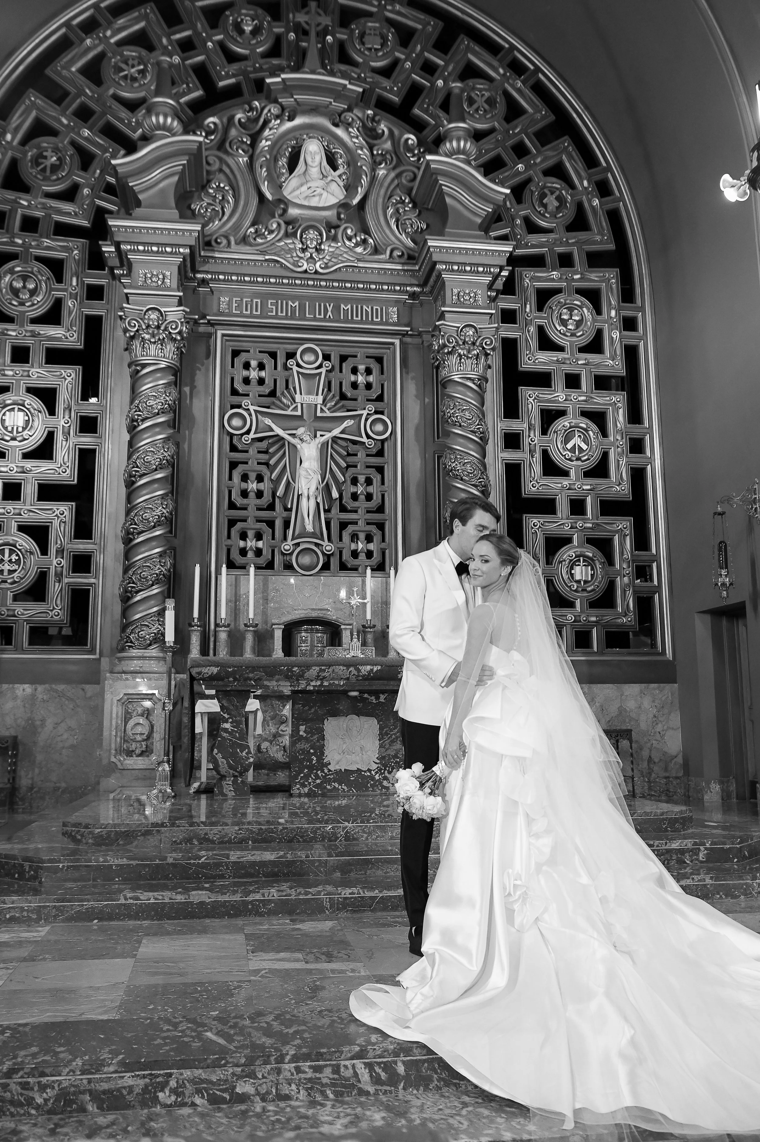 A bride and groom standing inside a church, holding hands, with an ornate altar and religious symbols behind them, including a crucifix and statues.