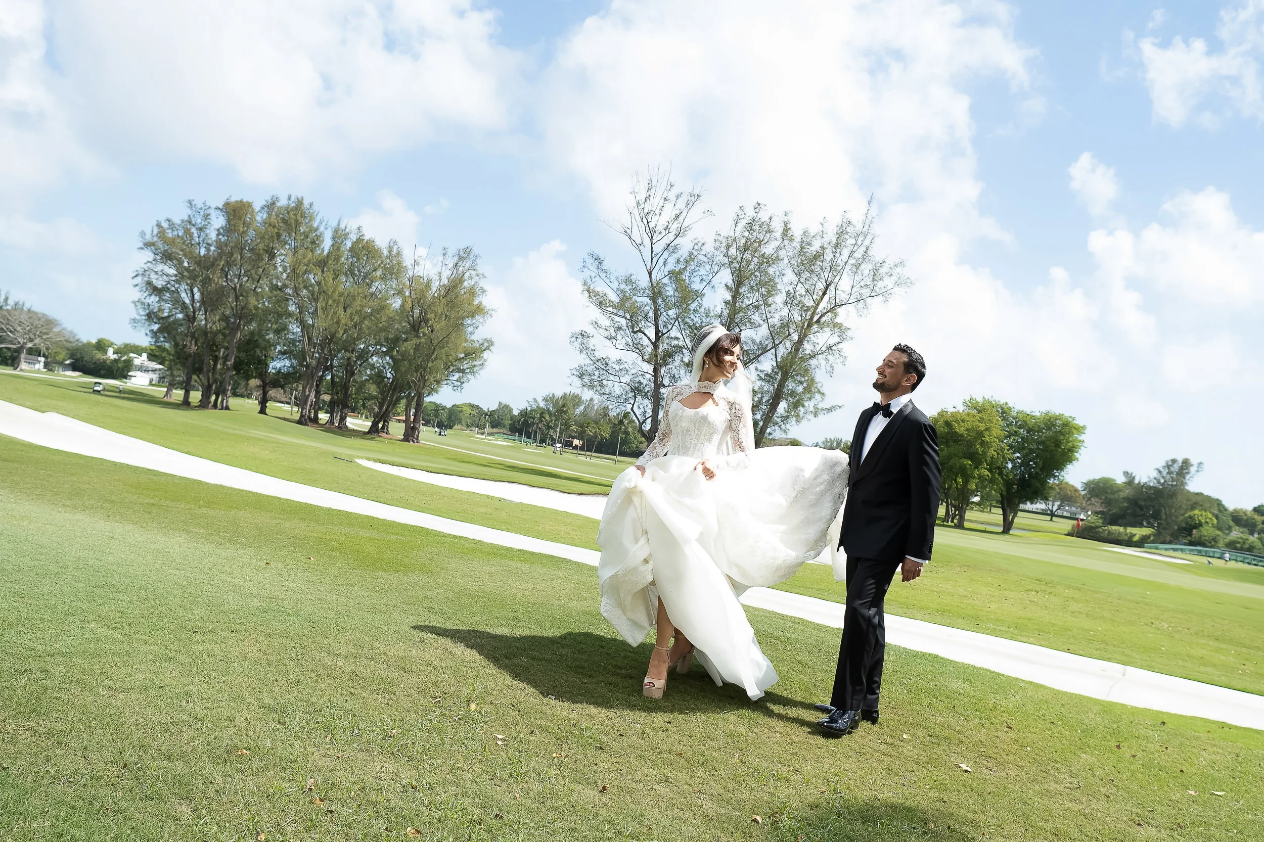 A bride and groom standing on a golf course on a sunny day with blue sky and clouds. The bride is in a white wedding dress and the groom is in a black tuxedo. The bride is holding her dress up slightly with one hand, and the groom is smiling at her.