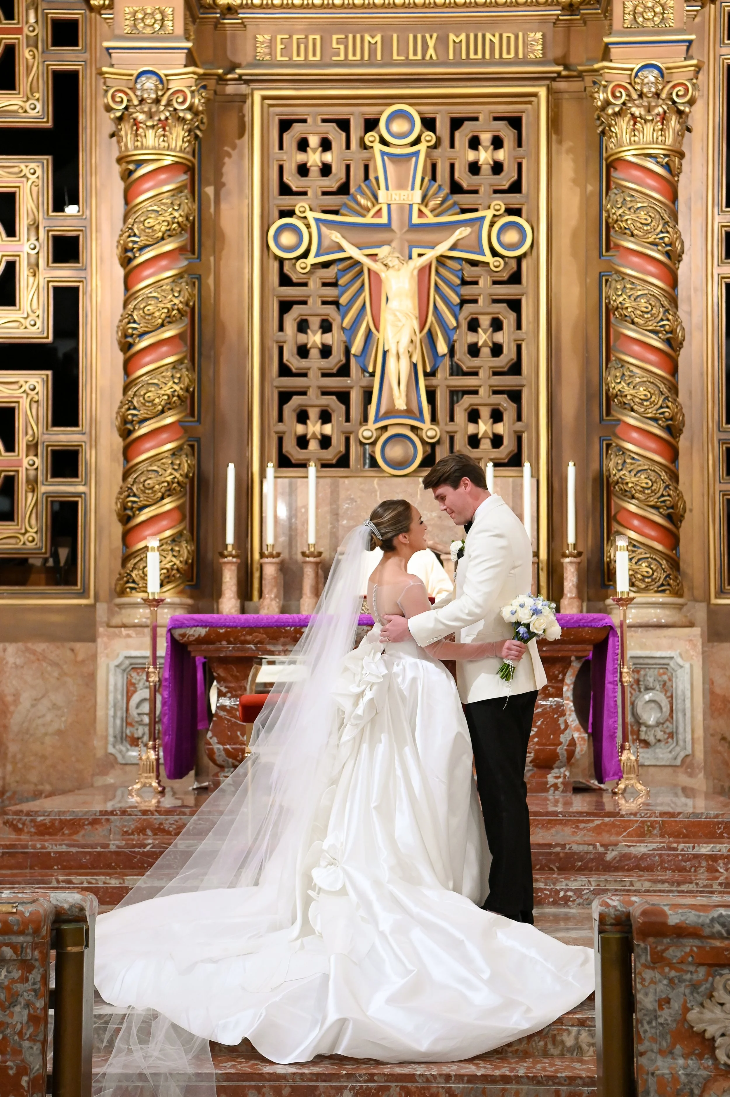 A bride and groom exchanging vows at an altar inside a church, with ornate gold and wood architectural details and a crucifix behind them.