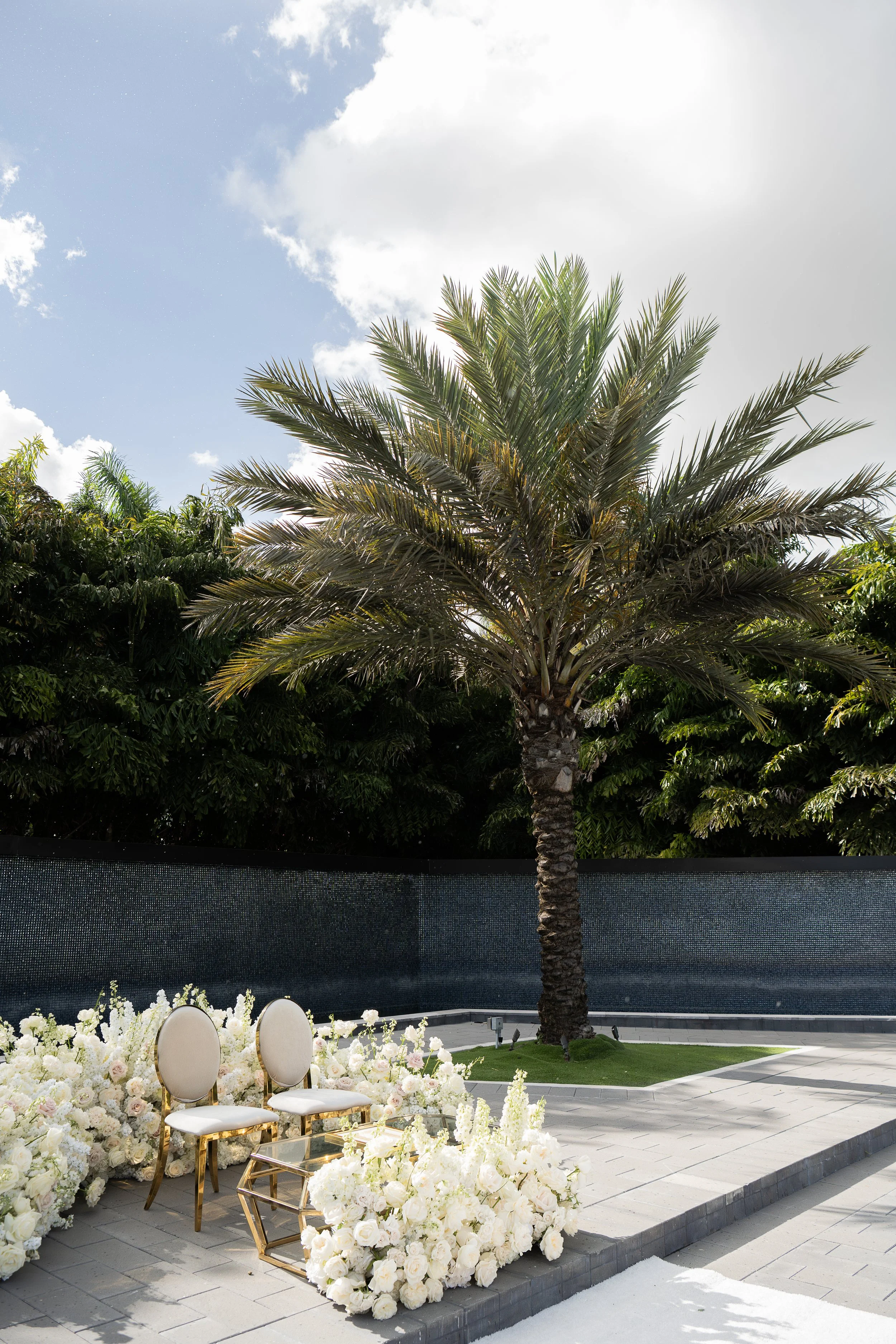 Outdoor event setup with white chairs and floral arrangements, decorated with white flowers and gold accents, under a palm tree with a partly cloudy sky.