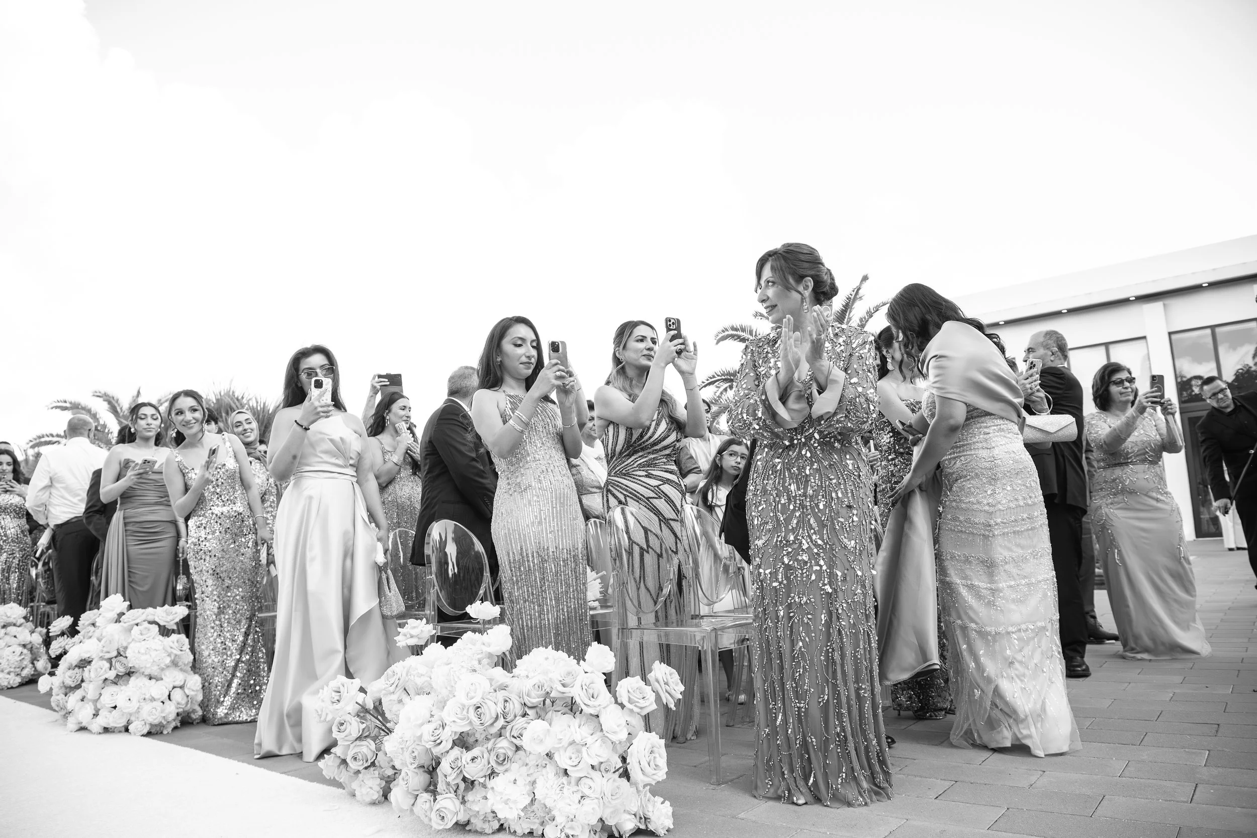 A black and white photo of a wedding ceremony with women dressed in elegant gowns standing behind a decorated floral arrangement, with some women taking photos and a woman wearing a detailed gown speaking to another woman in a wedding dress.