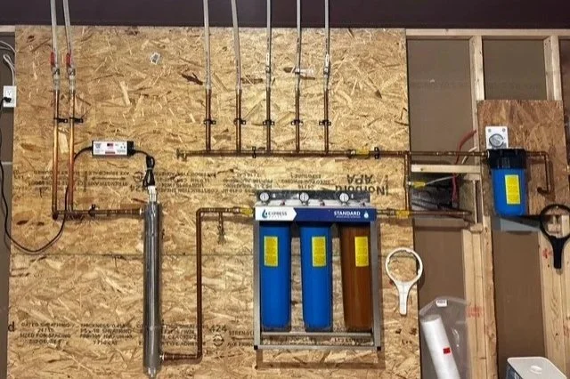 Plumbing and water filtration system mounted on a plywood wall, including pipes, valves, filters, and a wrench.
