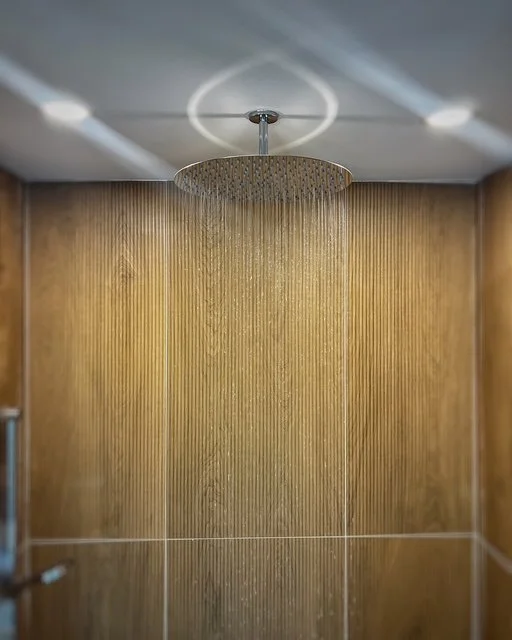 Modern shower with a ceiling-mounted rainfall showerhead, water cascading down, and a wood-paneled wall background.