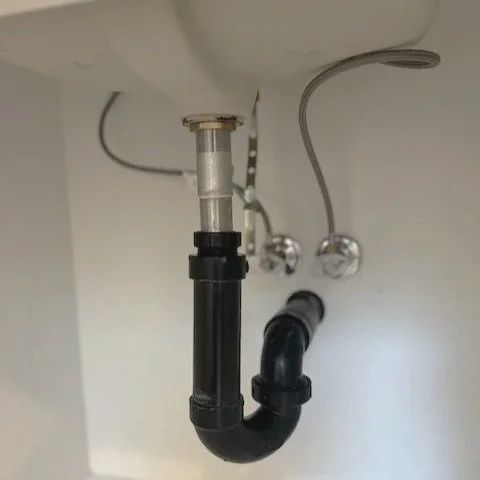 Underneath a kitchen sink, showing a black P-trap pipe connected to the drain. Two flexible water supply lines are visible above, attached to the faucet and the wall.