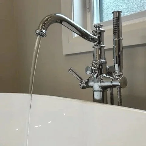Water flowing from a bathroom faucet into a tub with a window in the background.