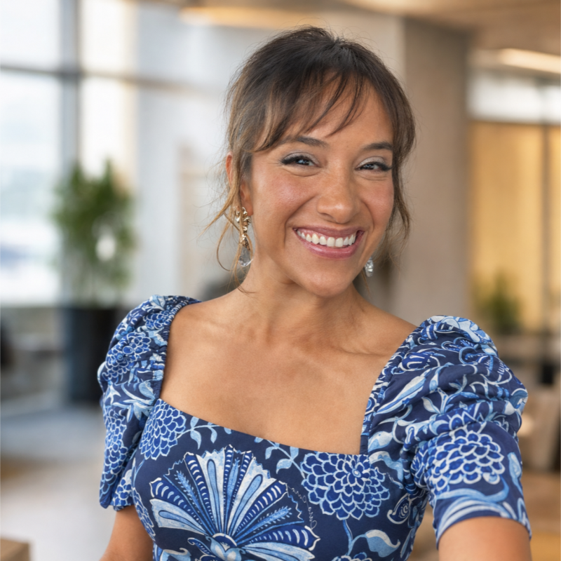 A woman with brown hair in a casual updo, wearing a blue patterned dress, smiling warmly in a modern, well-lit indoor space with plants in the background.