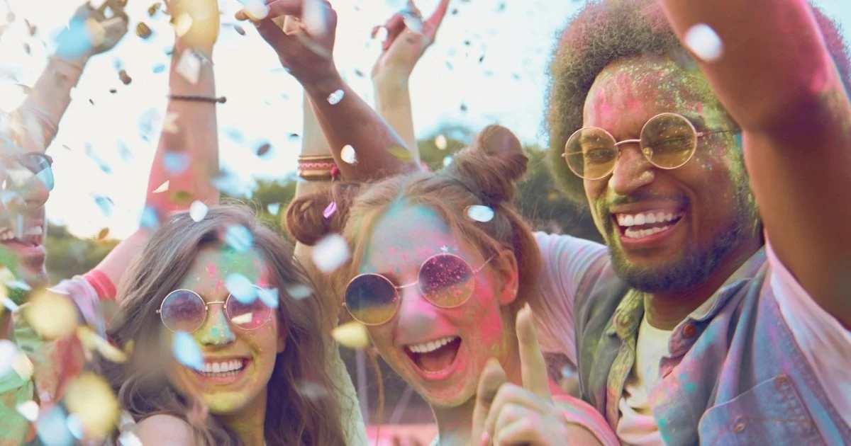 Group of friends celebrating with colored powder and confetti at an outdoor festival, smiling and wearing sunglasses.