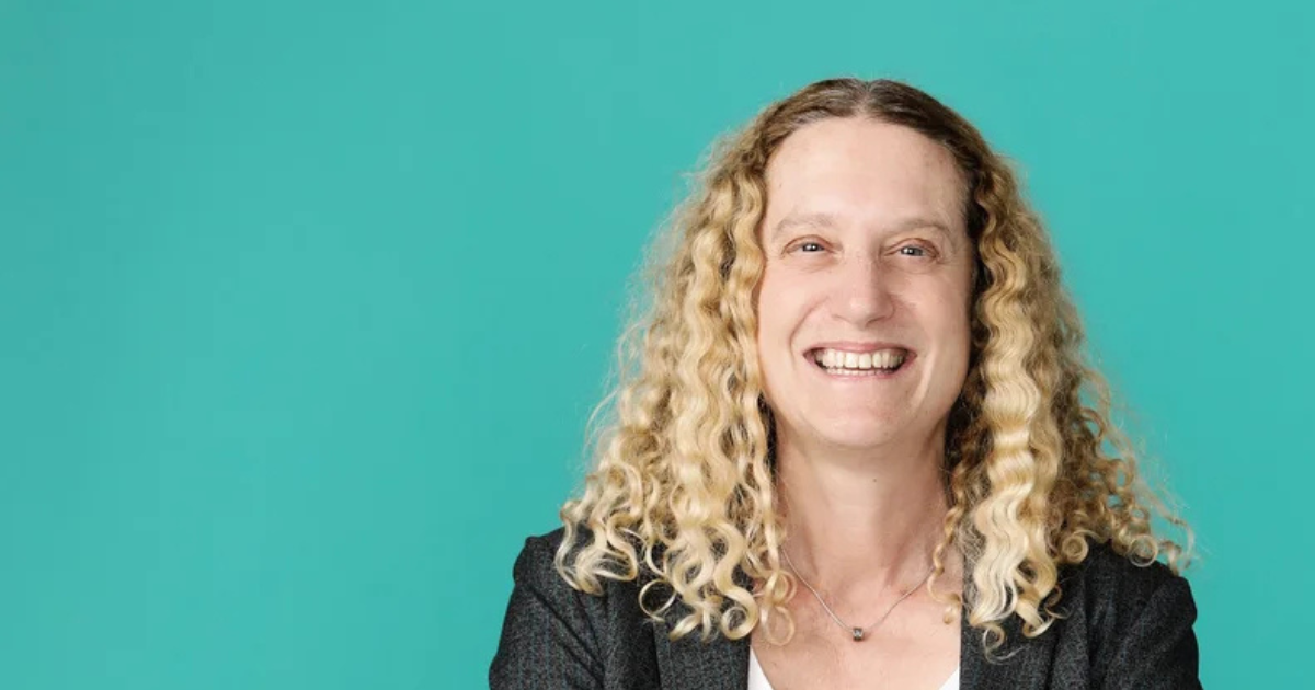 A woman with curly blonde hair smiling against a teal background.