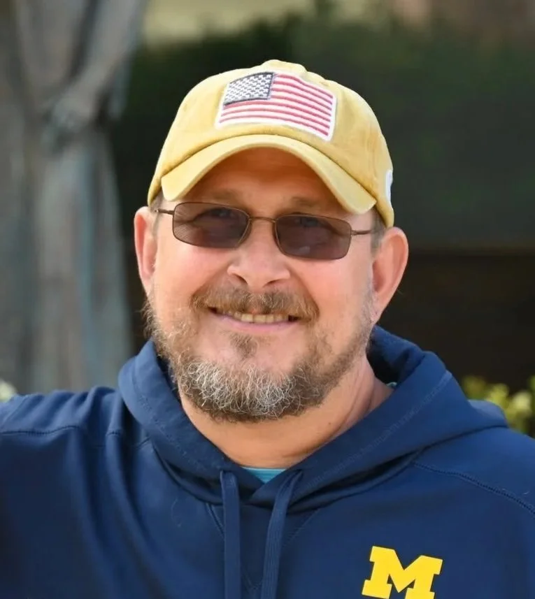 A smiling man wearing sunglasses, a yellow cap with an American flag patch, and a navy hoodie with a yellow 'M' logo.