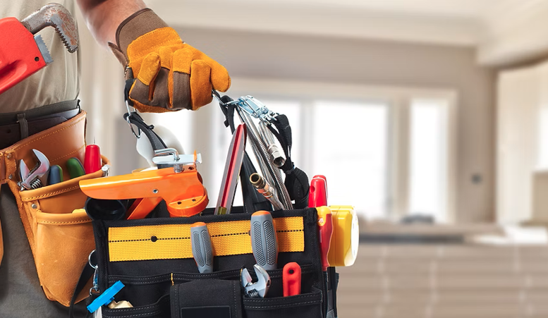 Person wearing work gloves holding a tool belt filled with various tools and hardware items in a room with natural light.