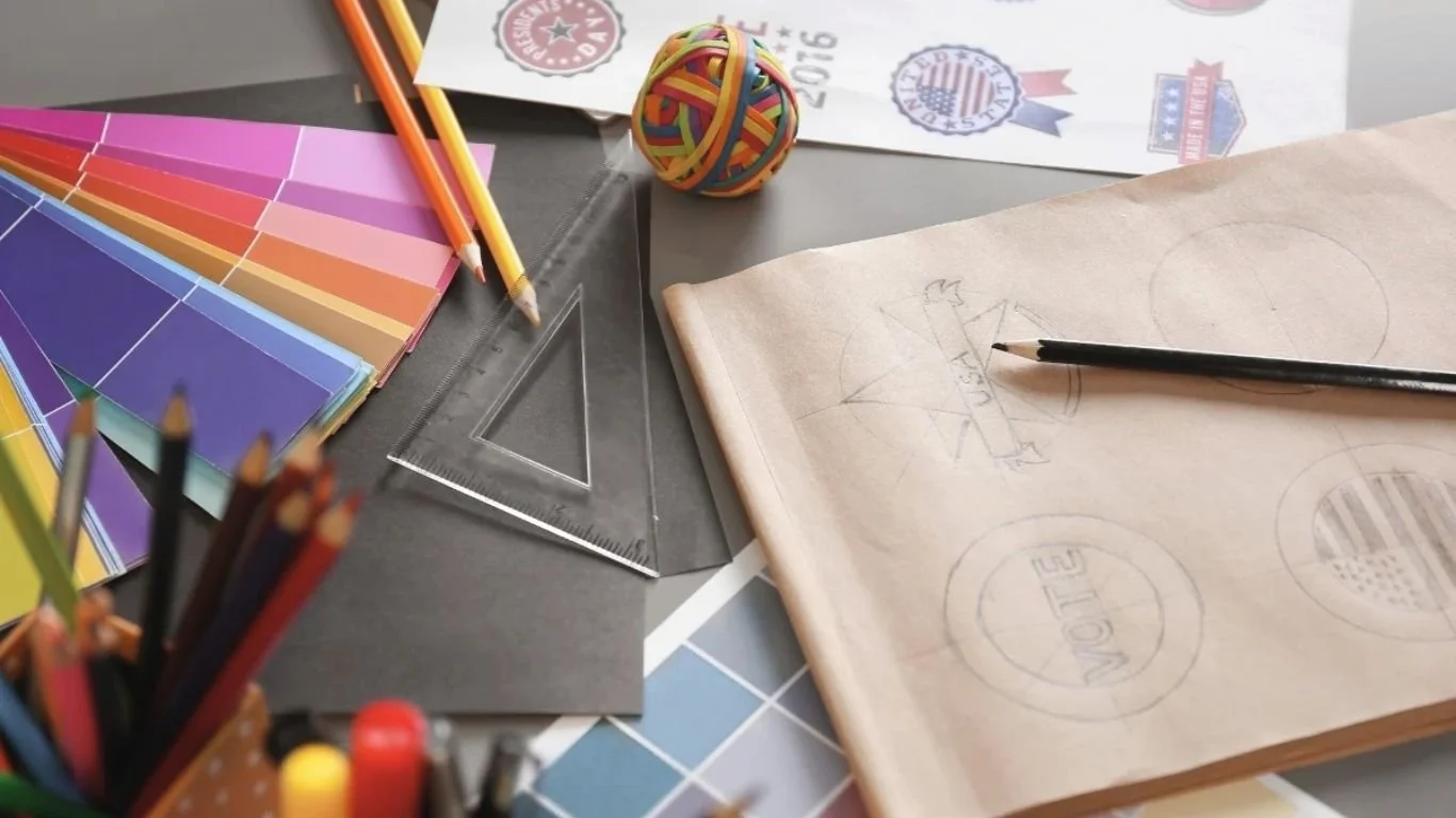 A sketchbook with pencil drawings of a star and the word VOTE sits among drafting supplies for graphic design, including a triangle ruler, colored pencils, and a ball of rubber bands.