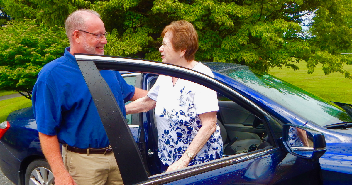 A man and a woman are smiling and talking while standing next to a blue car, with the woman inside the car and the man outside the car. The scene takes place outdoors in a park or yard with green trees in the background.