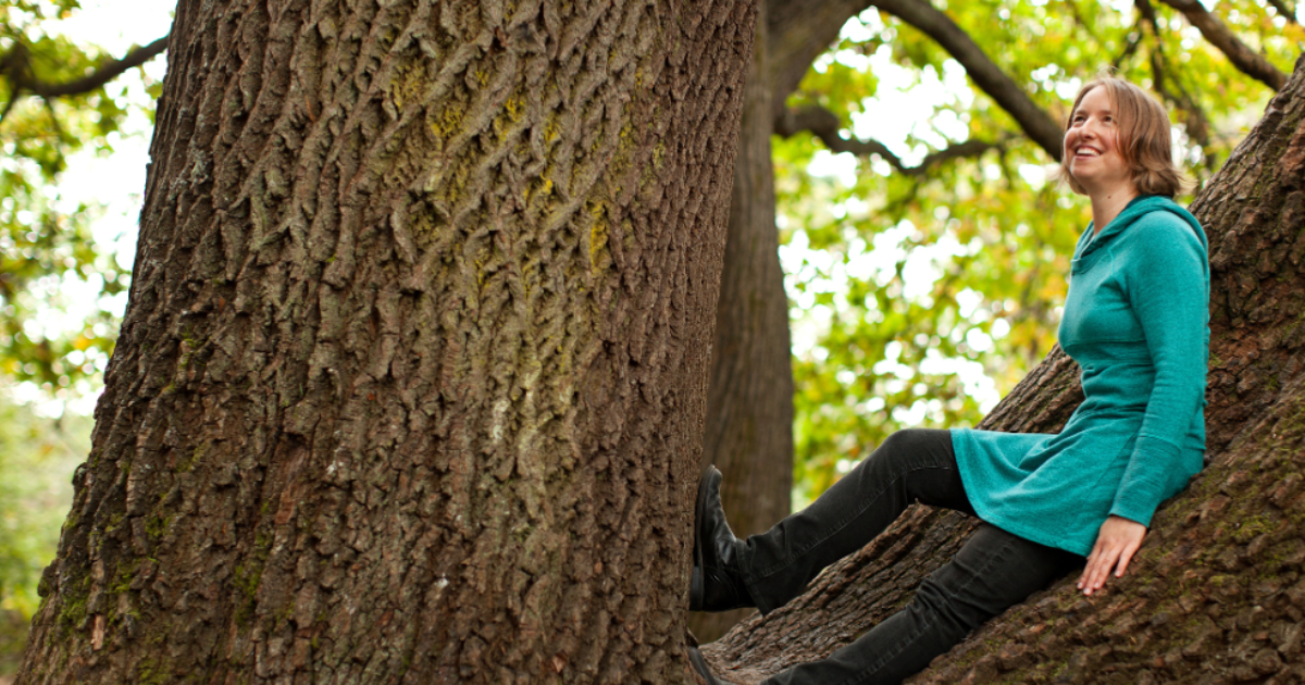 A woman with short brown hair smiling while sitting on a tree branch in a forest, wearing a teal hooded coat and black pants.