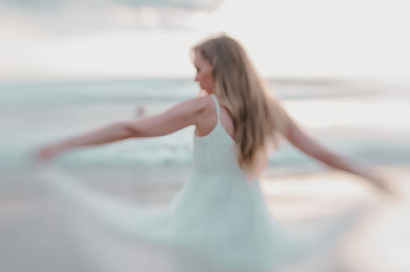 A woman in a white dress standing in shallow water at the beach, with arms extended and hair flowing in the breeze.