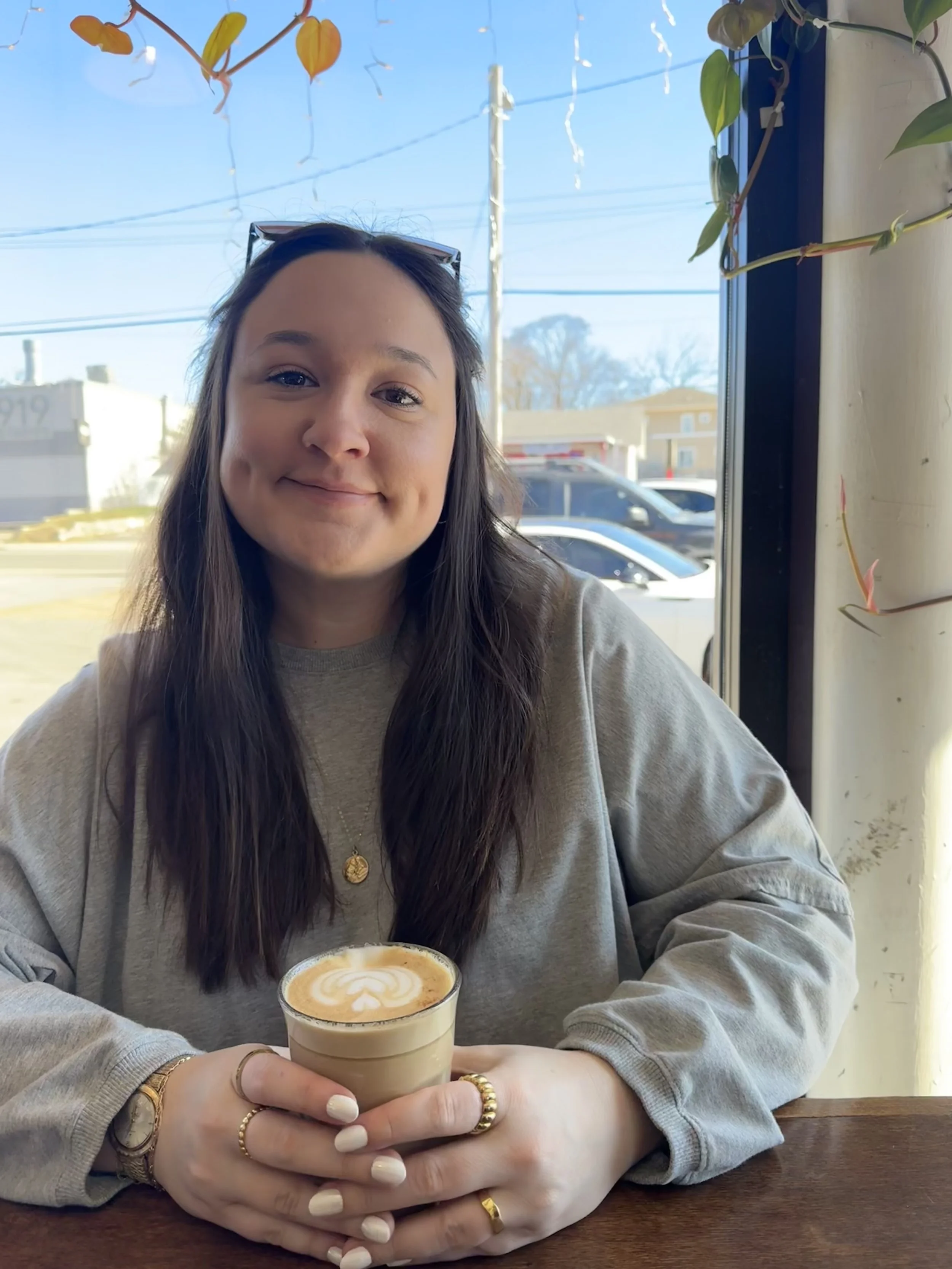 A young woman with long brown hair, wearing a grey sweatshirt, sitting at a wooden table in a cafe, holding a latte with latte art. She has sunglasses on her head and jewelry including rings, a bracelet, and a necklace. There are hanging plants and a window behind her, with cars and buildings visible outside.