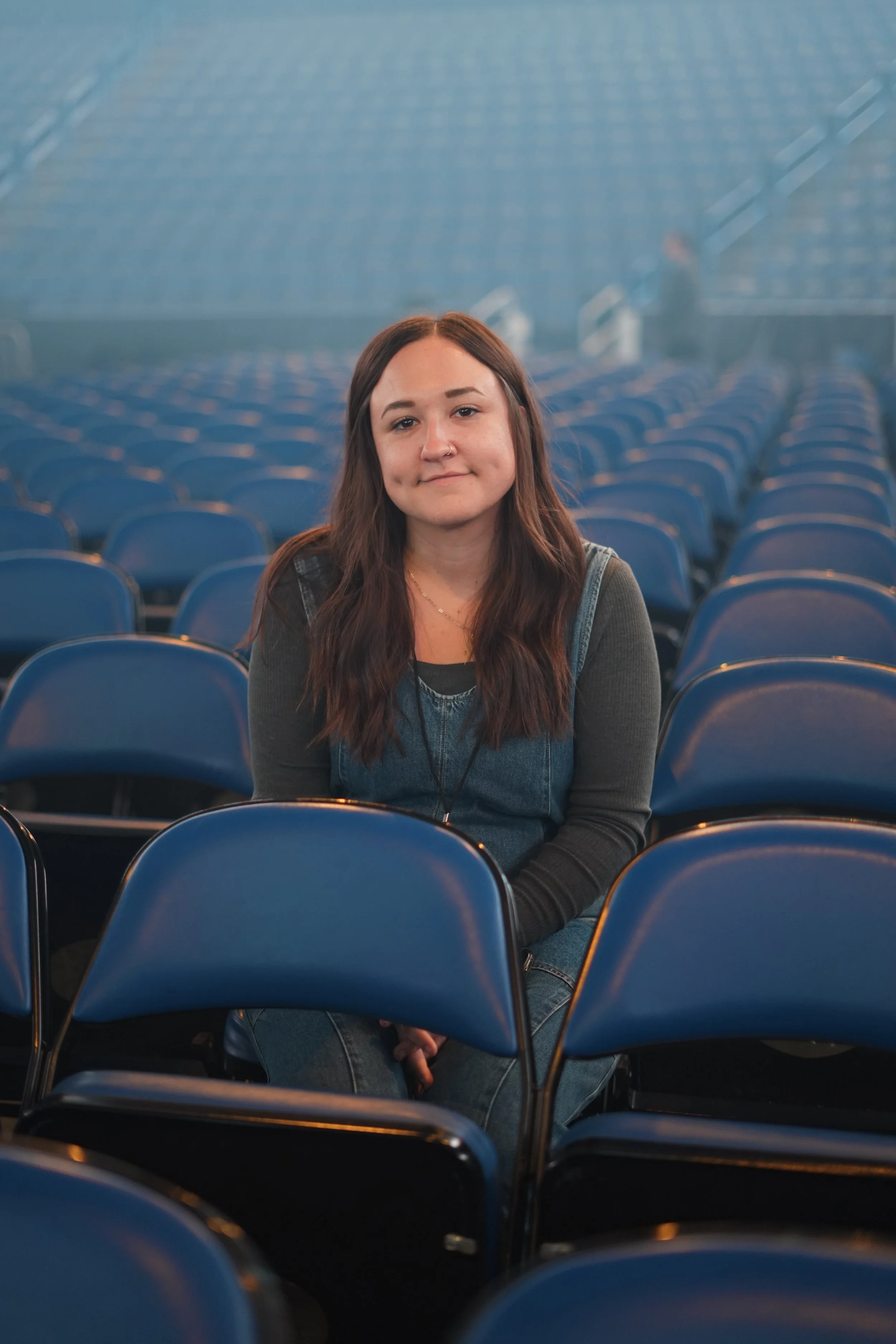 A young woman with long brown hair sitting among rows of blue folding chairs in an auditorium or theater, smiling slightly at the camera with a few people in the background.
