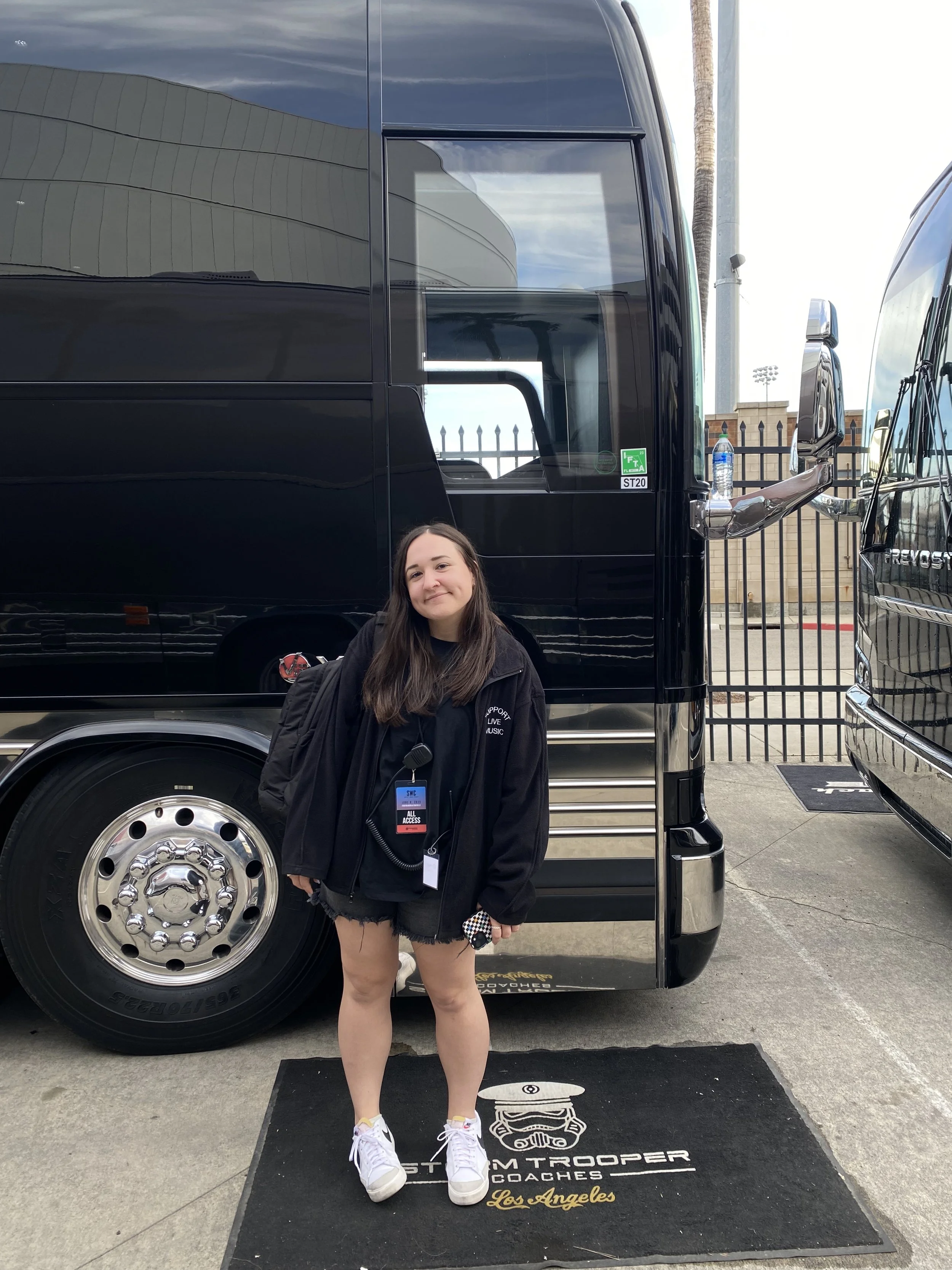 A young woman standing in front of a black bus, smiling. The bus has a reflective surface, and there is a water bottle on the bus's side mirror. The background includes a fence, a lamppost, and part of another bus.