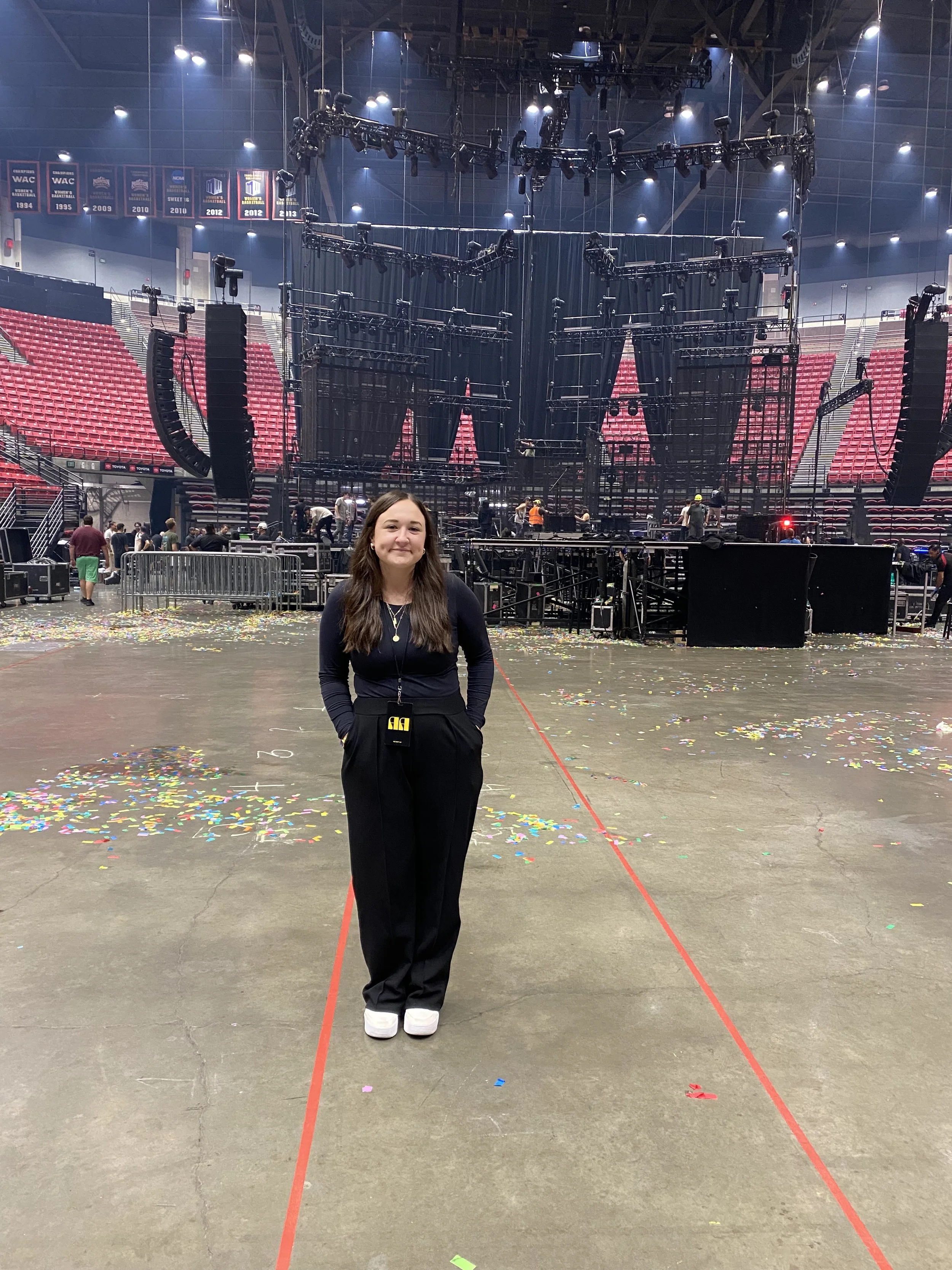 A young woman stands on a large indoor arena floor with a stage and many crew members preparing for an event. Colorful confetti is scattered on the floor, and red lines are marked on the ground.
