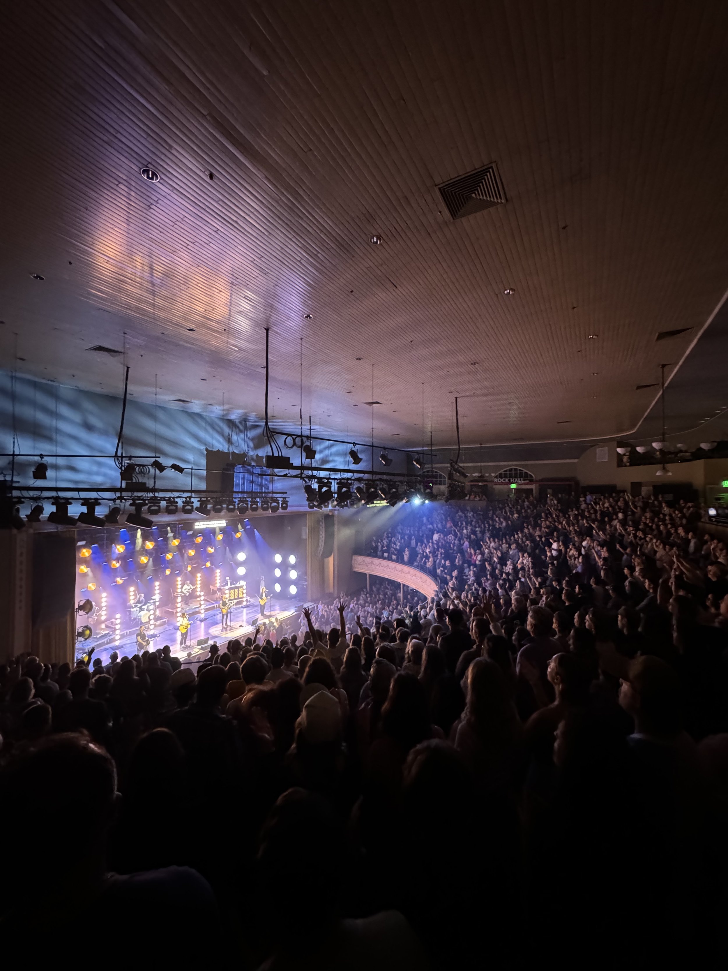 Indoor concert with a large audience, stage lit with bright lights, musicians performing with guitars, and a wooden ceiling overhead.