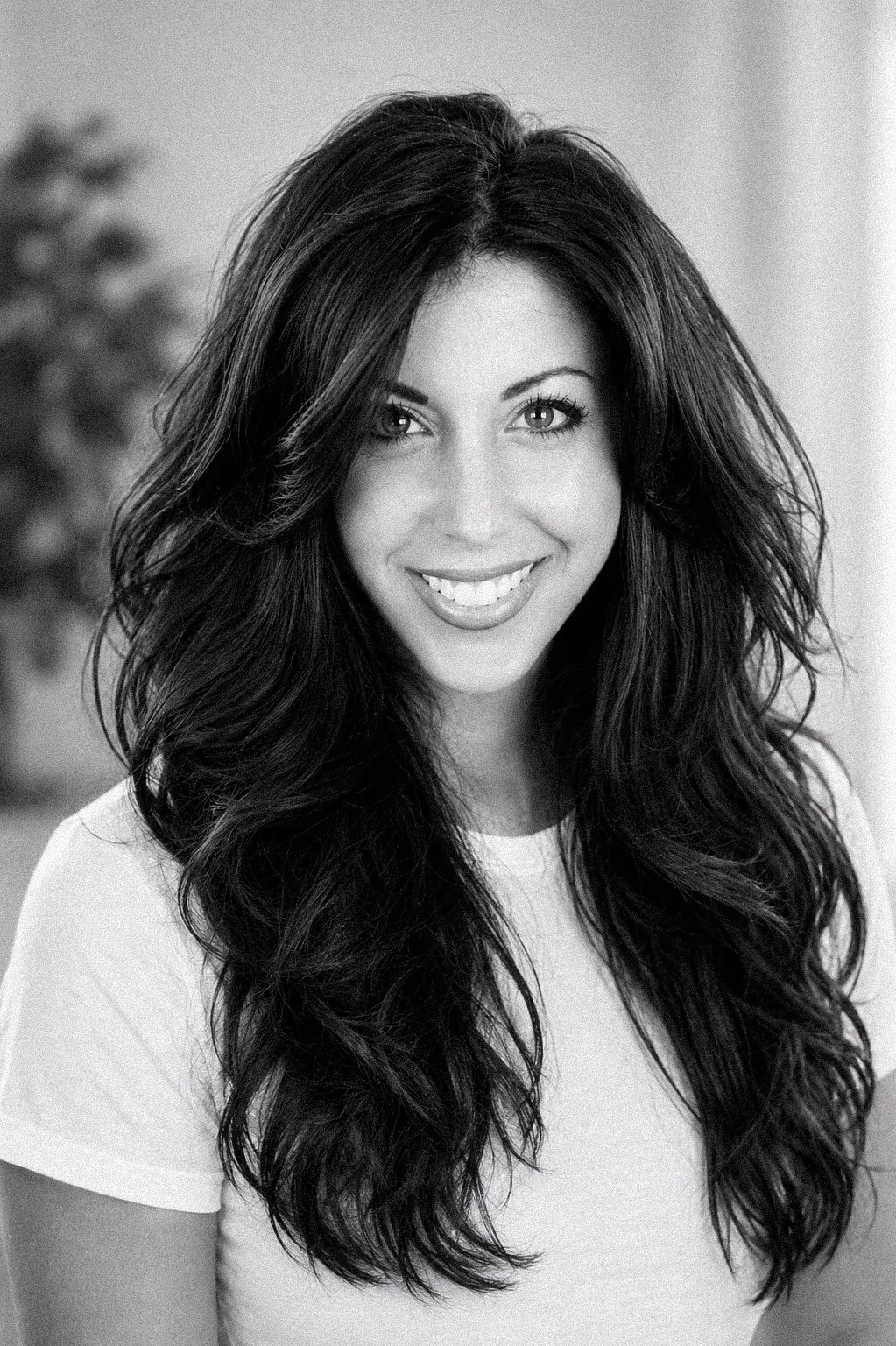 Black and white portrait of a woman with long, wavy hair smiling at the camera, wearing a white shirt with a blurred background.