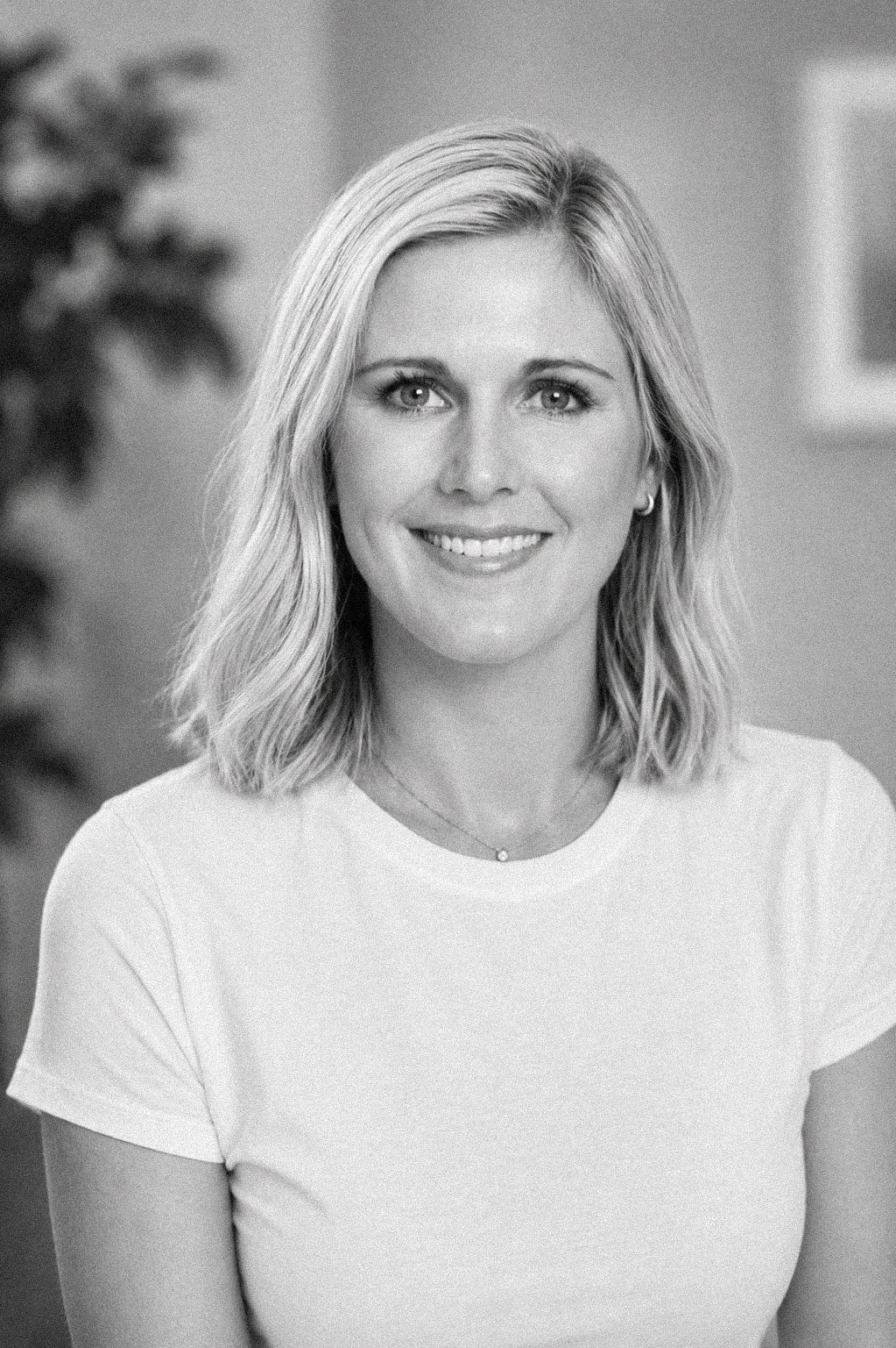 Black and white photo of a woman with shoulder-length blonde hair, smiling, wearing a light-colored t-shirt and a small necklace, against a blurred indoor background.