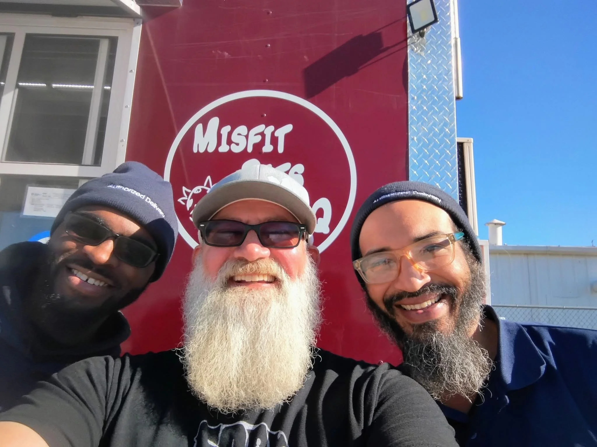 Three men taking a selfie in front of a red trailer with a logo reading "Misfit" and a cartoon graphic of a cat. One man has a white beard, glasses, and wears a beige cap. The other two men wear dark sunglasses and beanies.