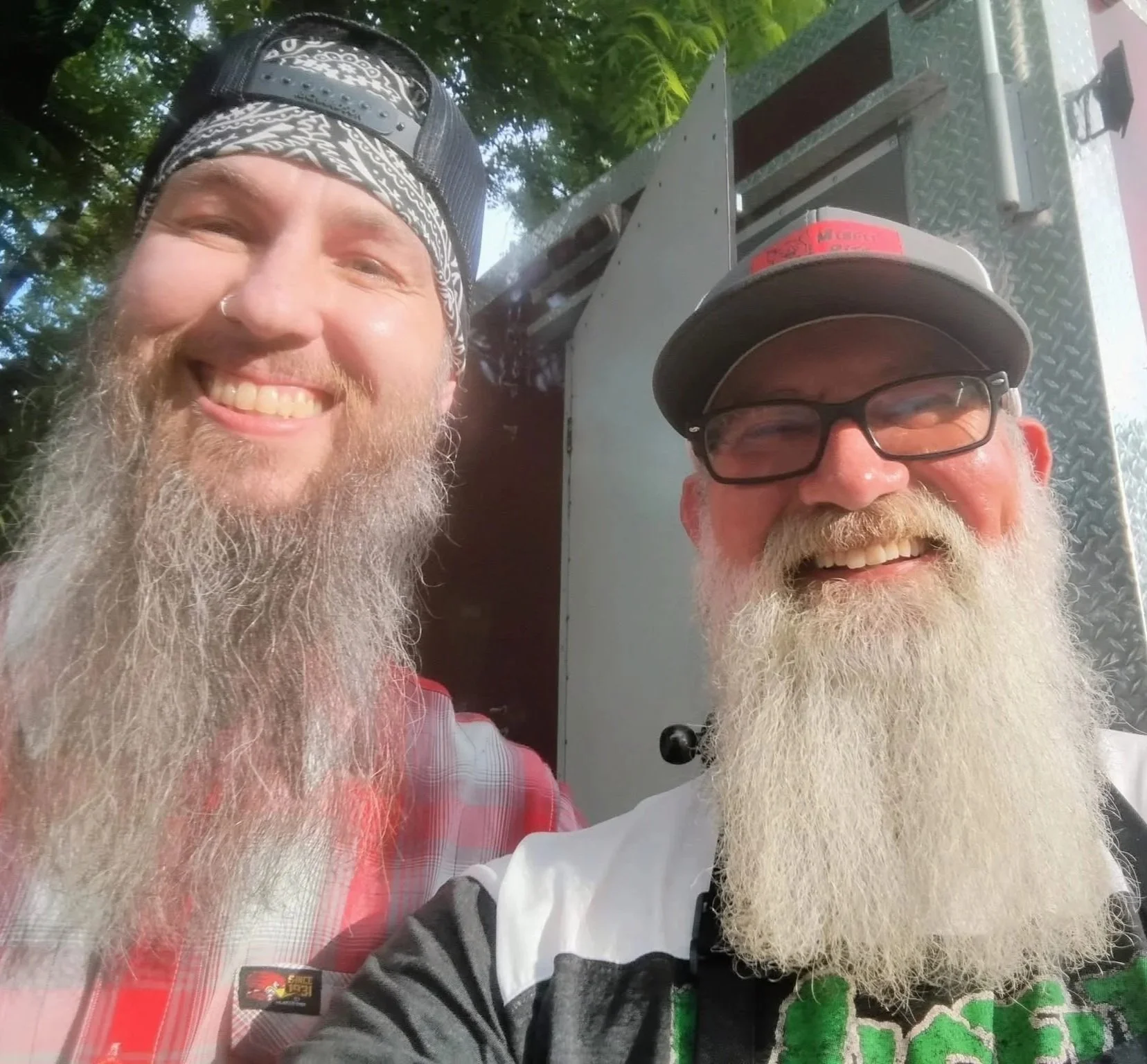 Two smiling men with long white beards and glasses, one wearing a baseball cap and the other wearing a cap and glasses, taking a selfie outdoors with greenery in the background.
