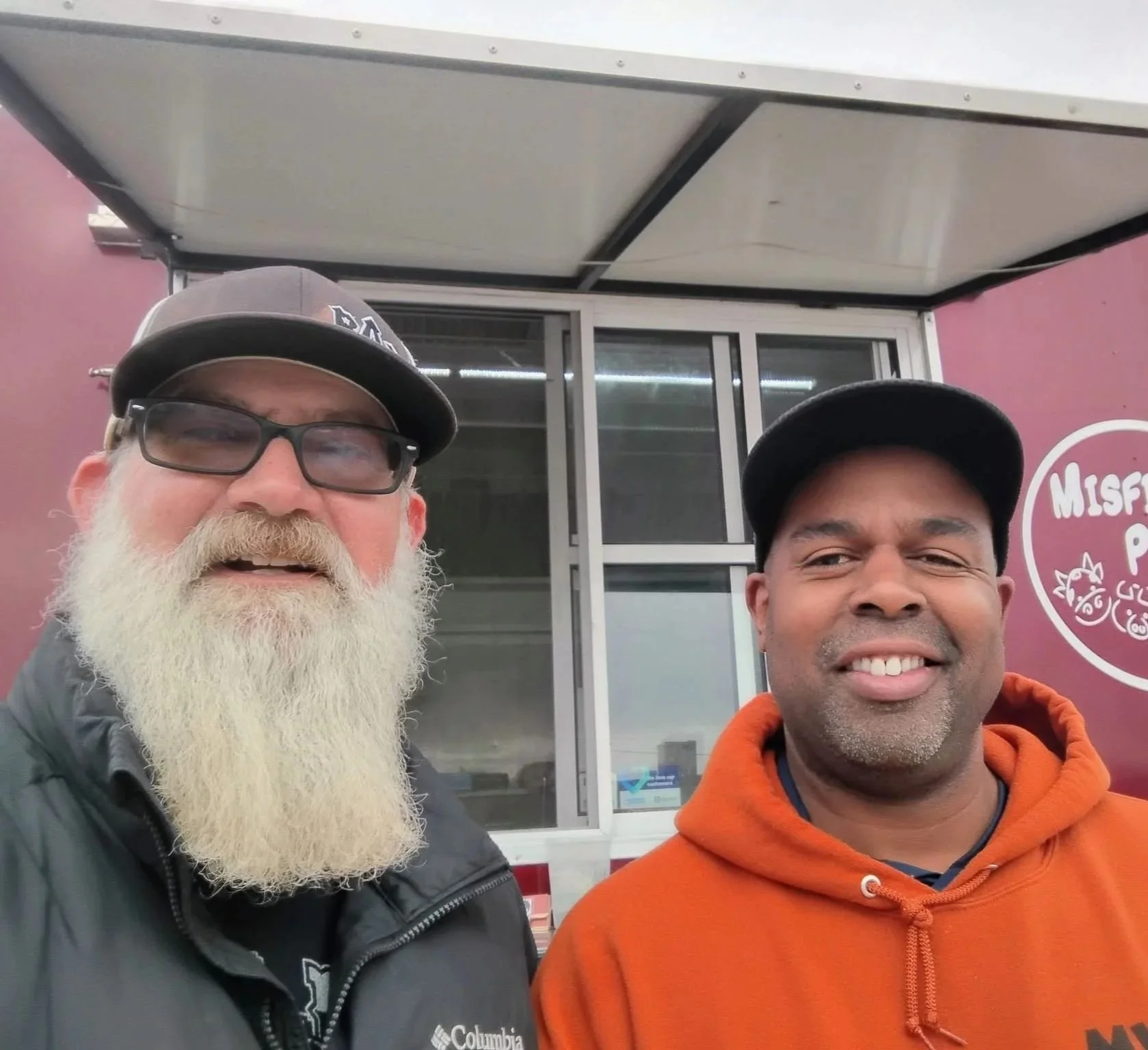 Two men taking a selfie in front of a purple building with a window and a partial logo visible.