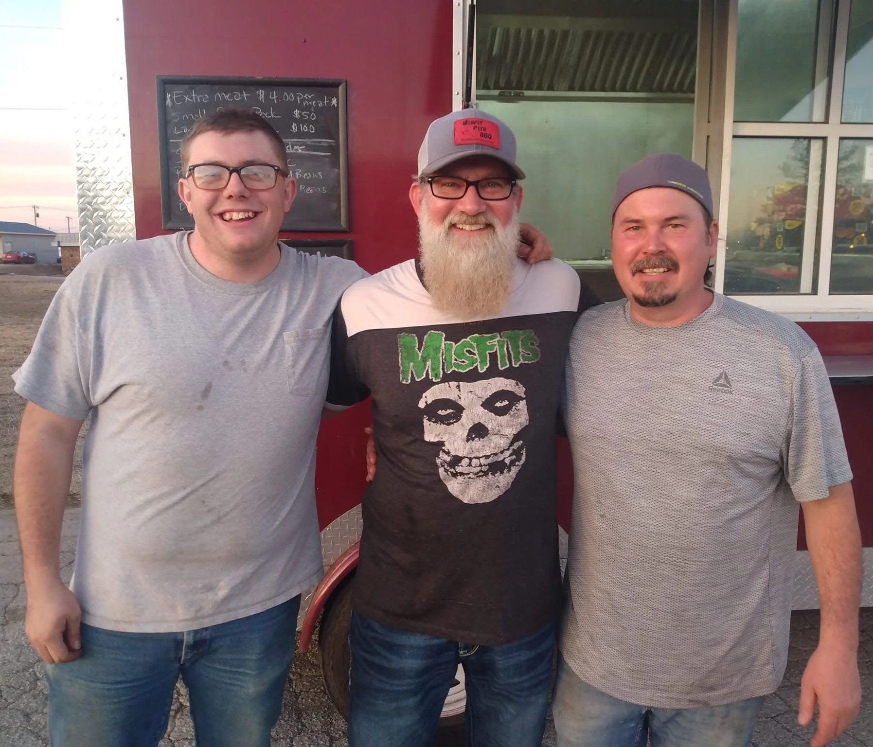 Three men with light skin and glasses smiling and standing close together outside food truck. The man in the middle has a long beard and wears a Misfits band t-shirt and a gray cap. The man on the left has short brown hair, wears a gray t-shirt, and 