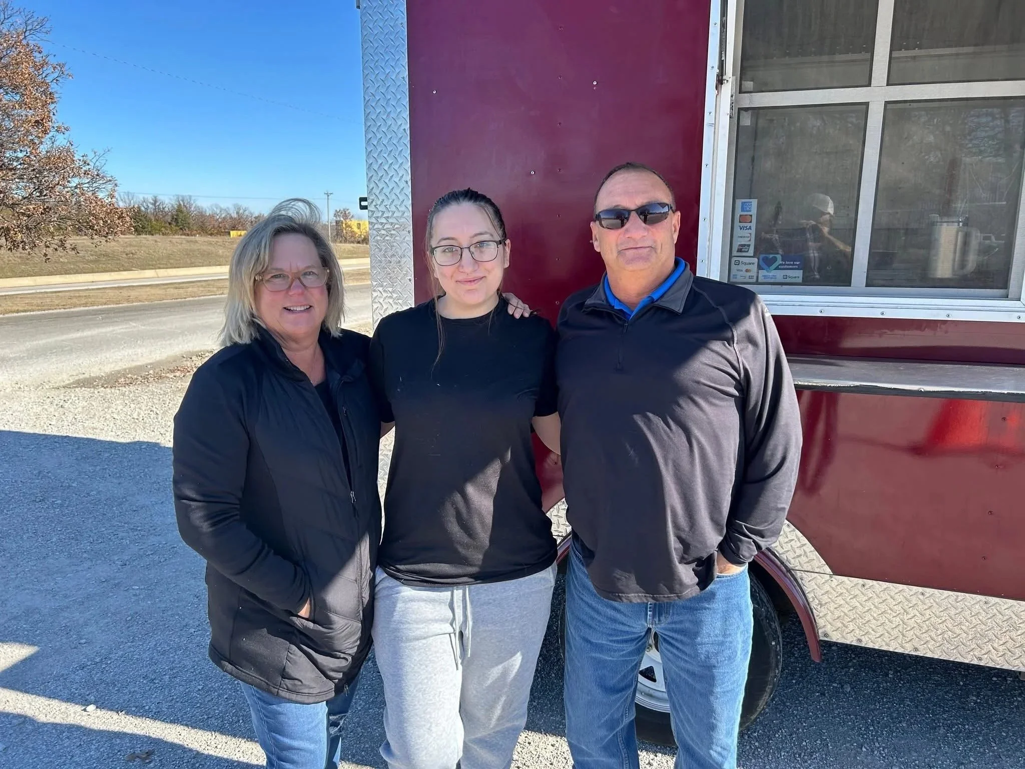 Three people outdoors standing in front of a red truck with a large window. It is a sunny day with clear blue sky. The group appears to be a family or friends, smiling and dressed casually.