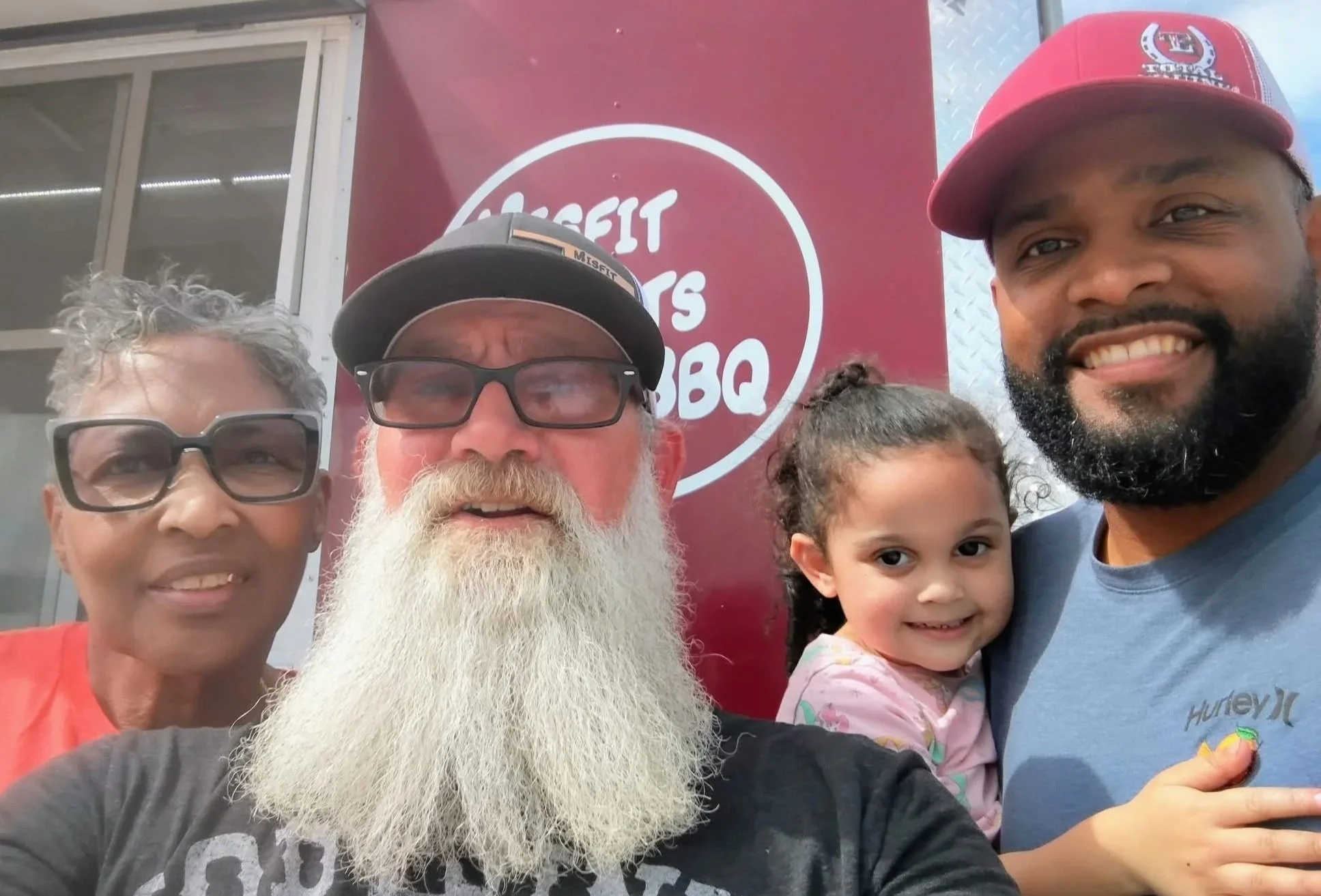 Family photo with five people smiling in front of a red sign with white text that reads 'Lageret Barbecue.'