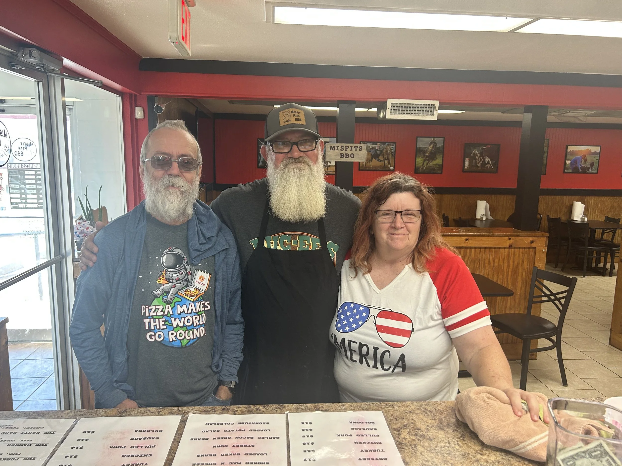 Three people standing together inside a restaurant, with framed pictures of horses and riders hanging on the wall behind them. The man on the left has a long gray beard and glasses, wearing a dark graphic t-shirt and a blue jacket. The man in the mid