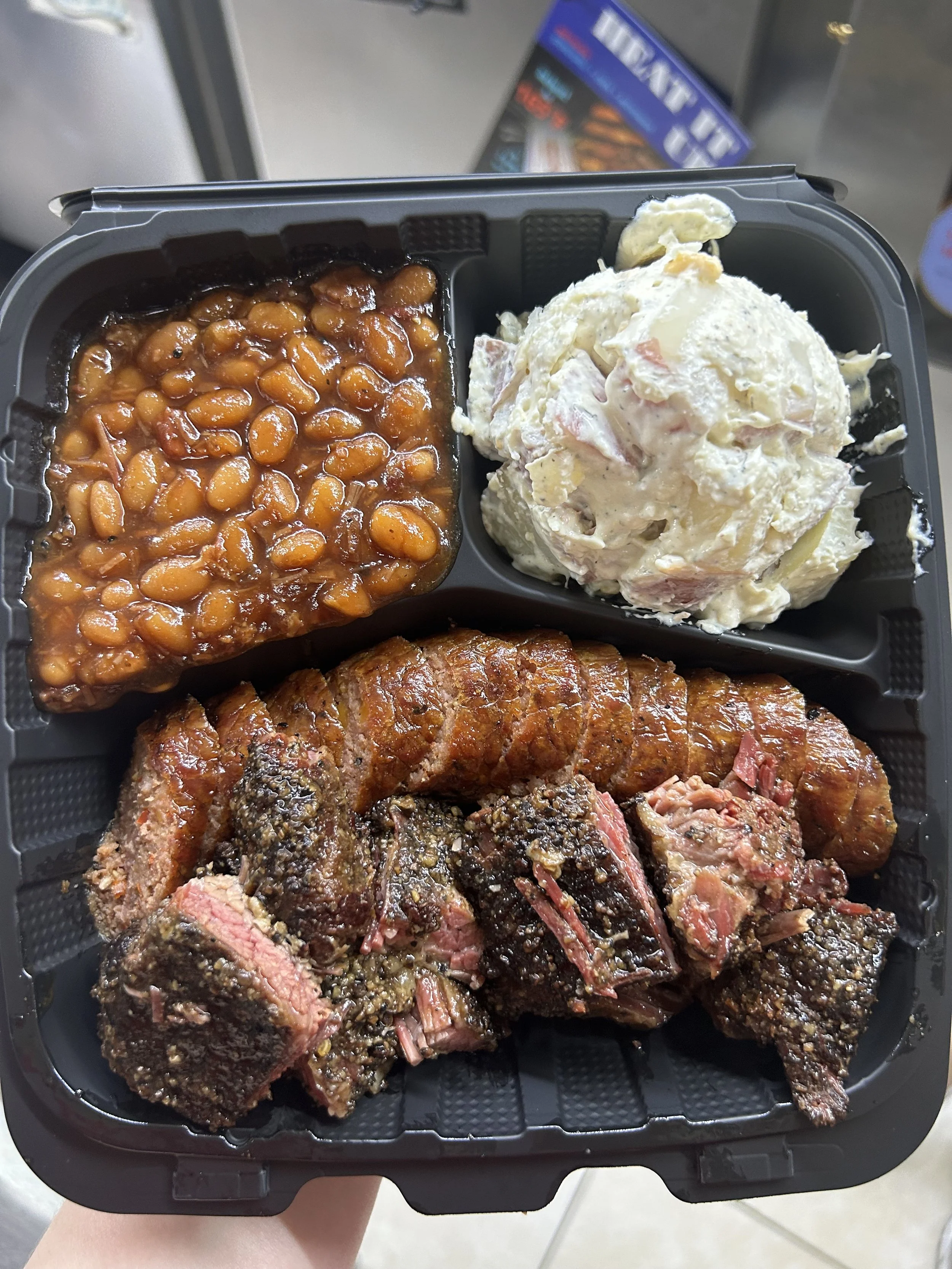 A black plastic tray containing brisket burnt ends, sliced hot sausage, a serving of baked beans, and a serving of potato salad.