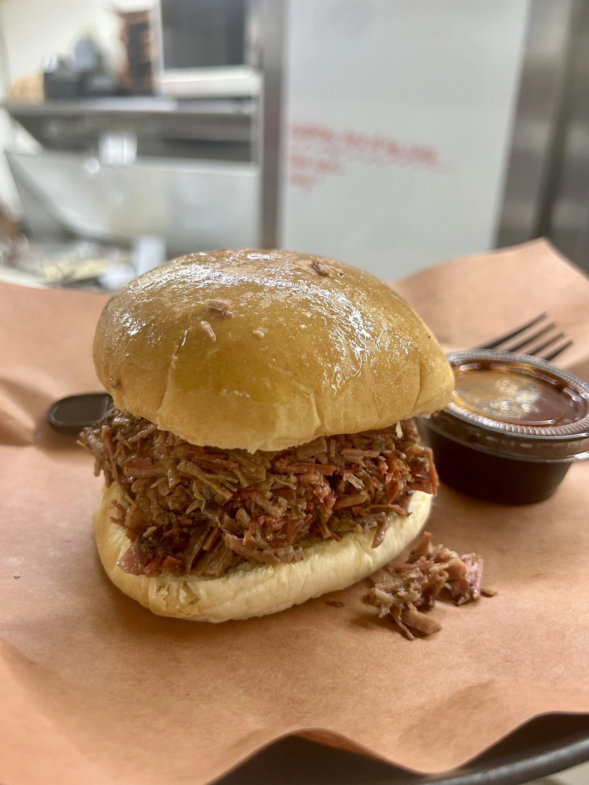 Chopped brisket sandwich with chopped brisket meat on a bun, served with a side of barbecue sauce in a small cup.