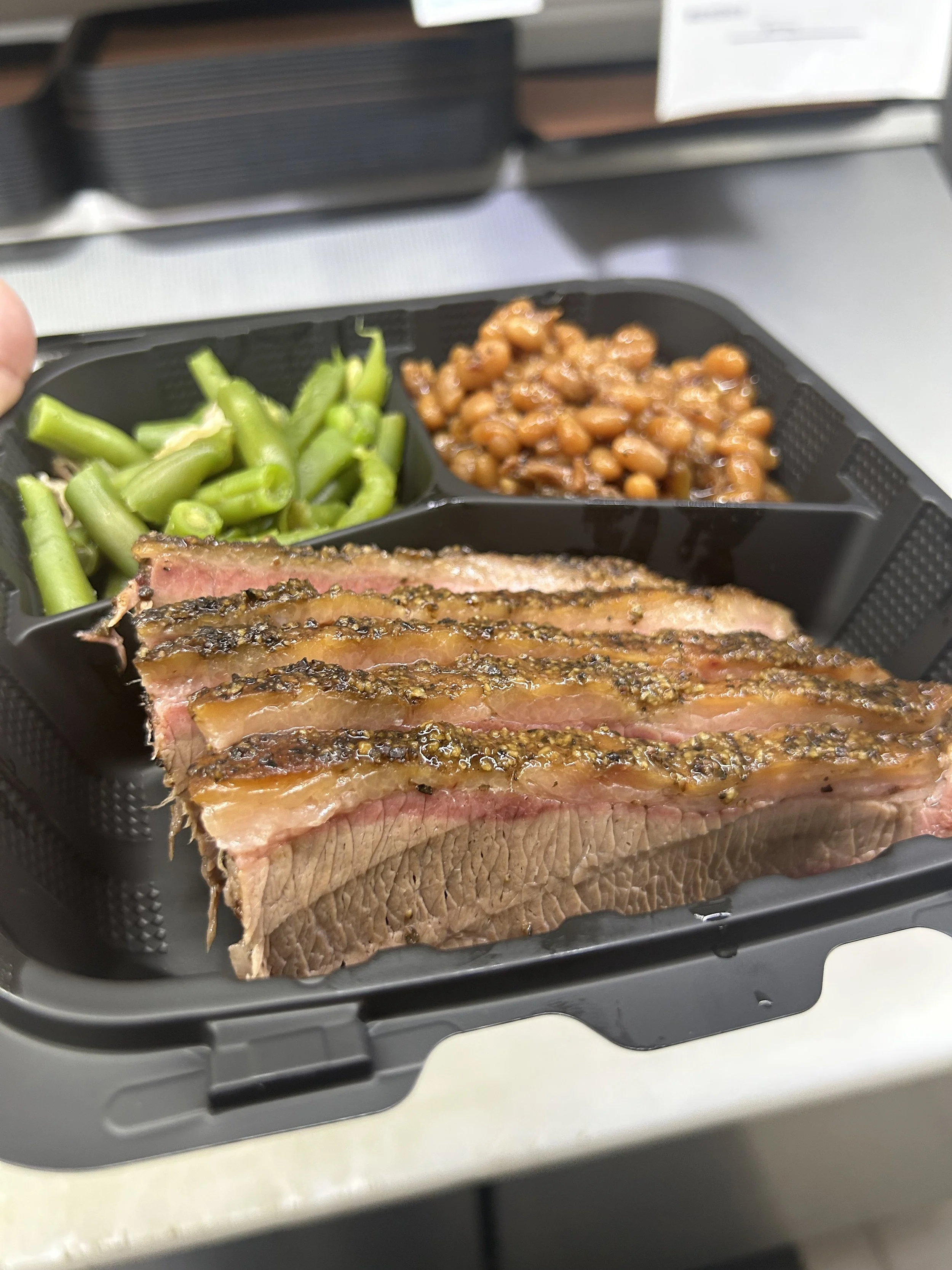 Cooked sliced beef steak with black pepper and herbs, green beans, baked beans, and rice in a black plastic food container.