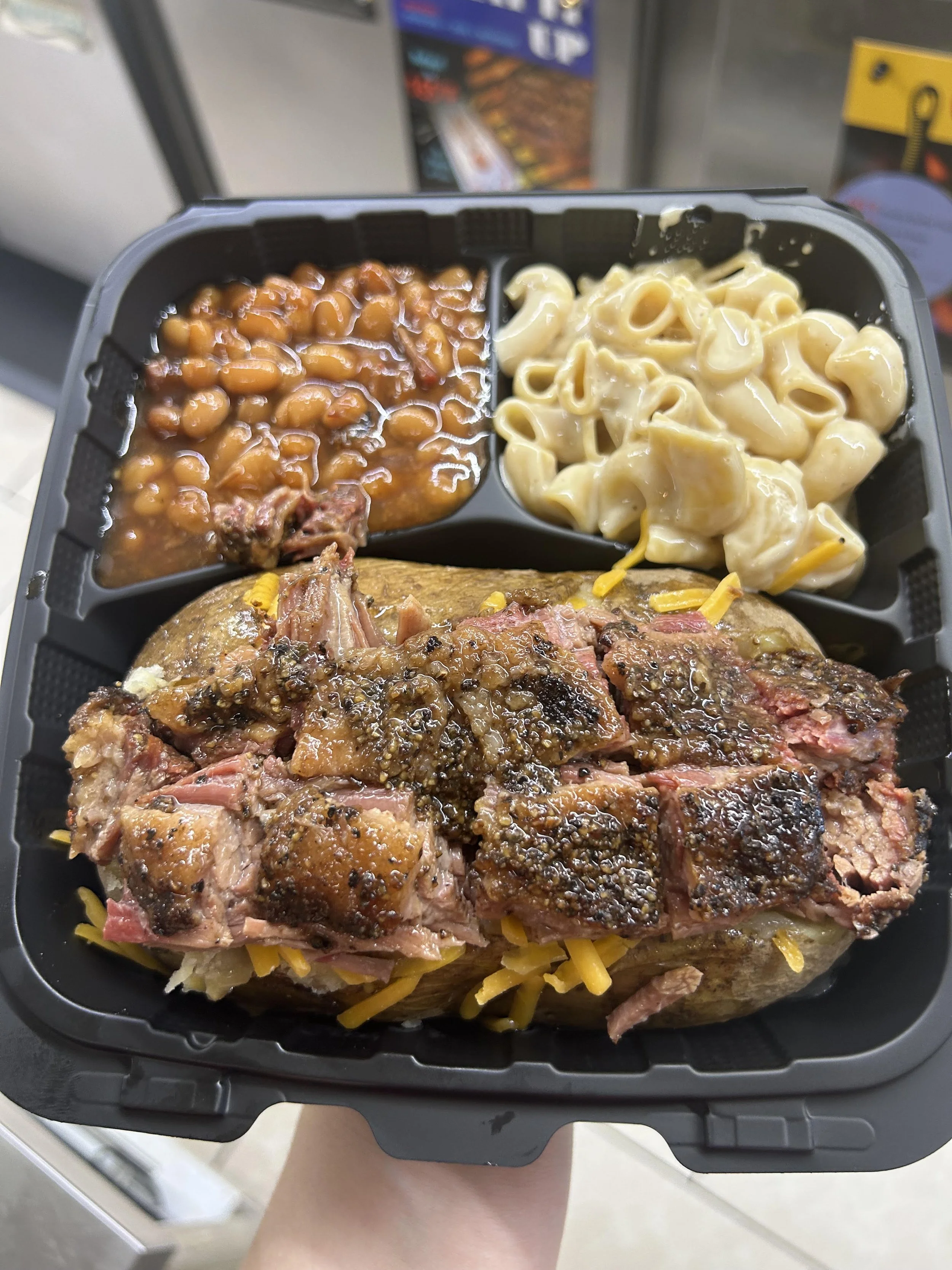 A black plastic tray with separated compartments containing baked beans in sauce, macaroni and cheese, and a baked potato with burnt ends.