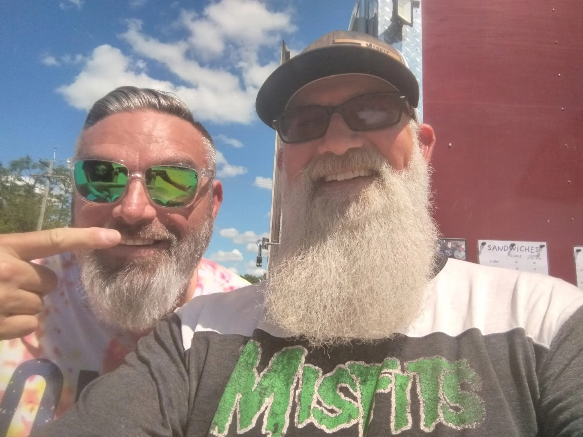 Two smiling men with beards and glasses taking a selfie outdoors on a sunny day, with a blue sky and clouds in the background. One man is wearing a baseball cap, sunglasses, and a t-shirt, while the other is wearing sunglasses and a colorful shirt.